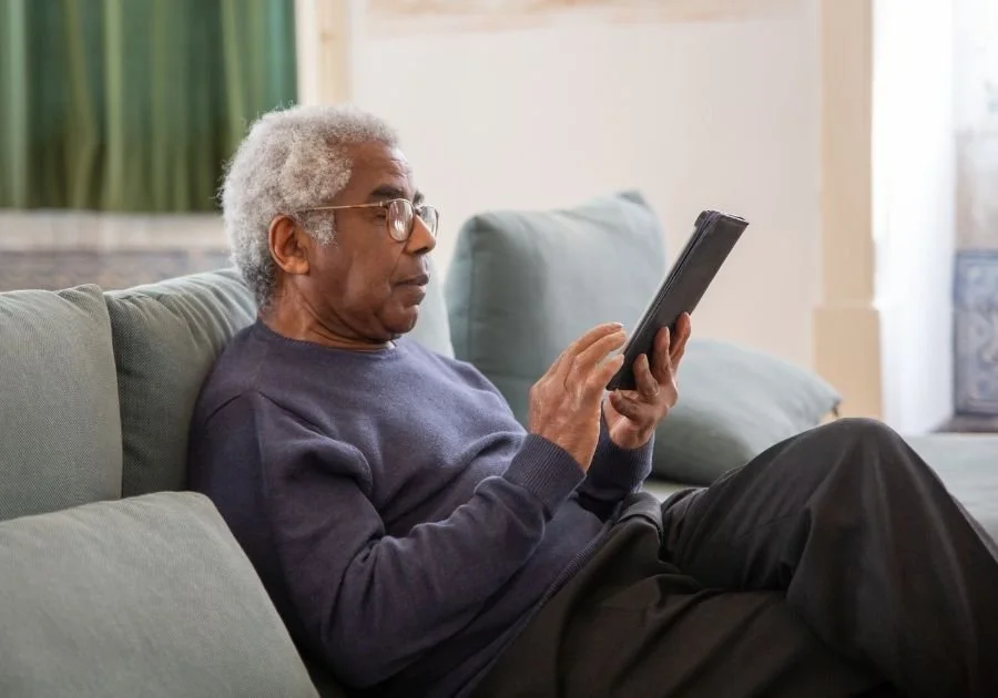 Older man with gray hair and glasses sitting on a light green couch, using a tablet in a bright living room.