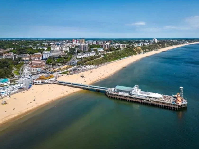 An aerial view of a pier extending into the ocean from a sandy beach, with a Ferris wheel and buildings visible on the shore, with Bournemouth town skyline in the background under a partly cloudy sky.