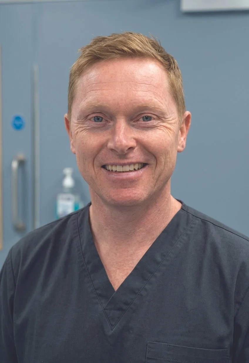 Our practitioner, Ryan McCormack, wearing medical scrubs, standing in a clinical setting with blue walls and medical equipment in the background.