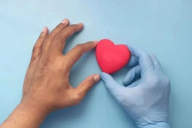 A person with a bare hand and a person wearing a blue medical glove are holding a red heart-shaped object on a light blue background.