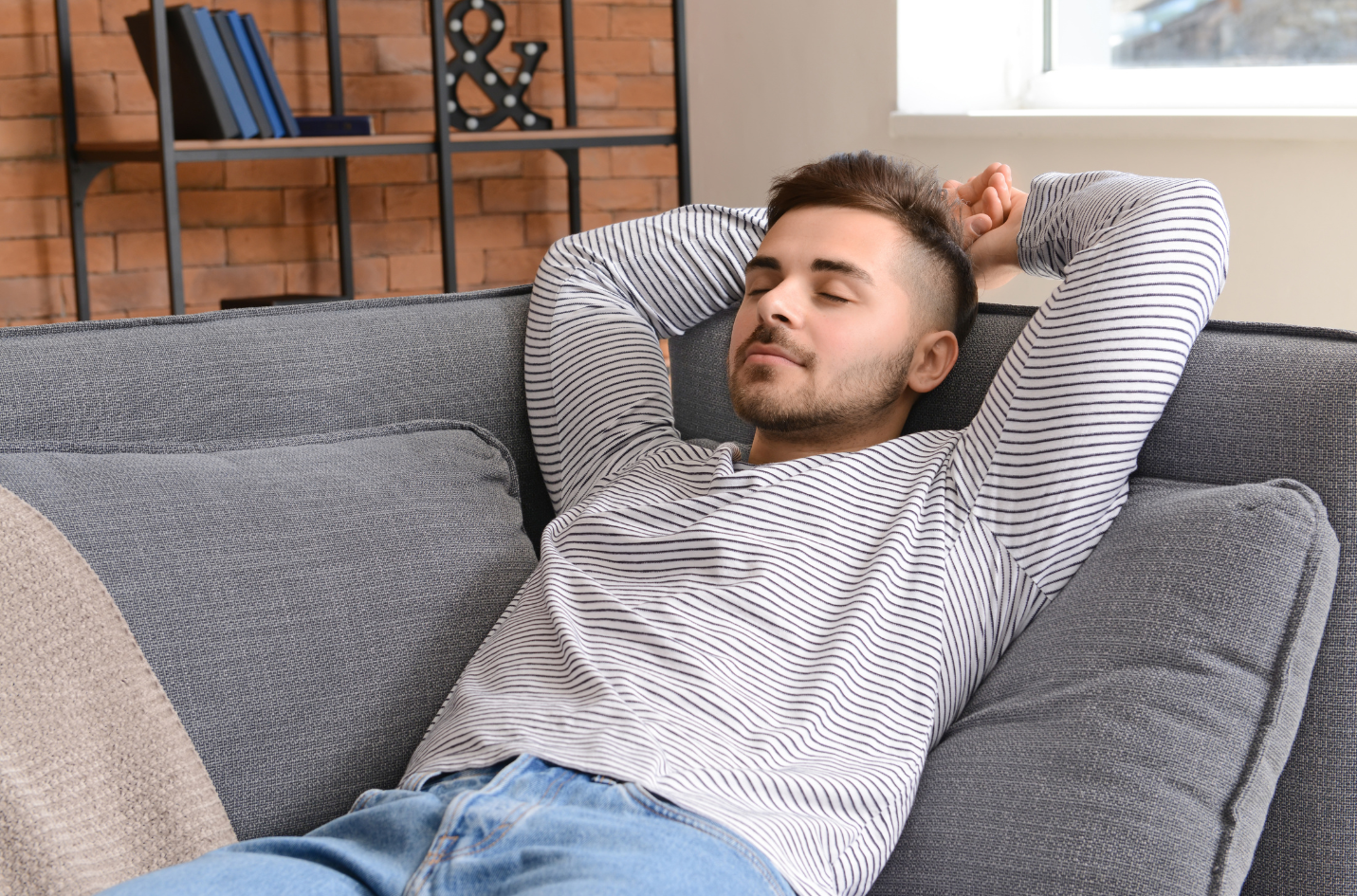 A person with arms behind their head rests on a gray sofa, eyes closed, wearing a striped long-sleeved shirt and blue jeans.