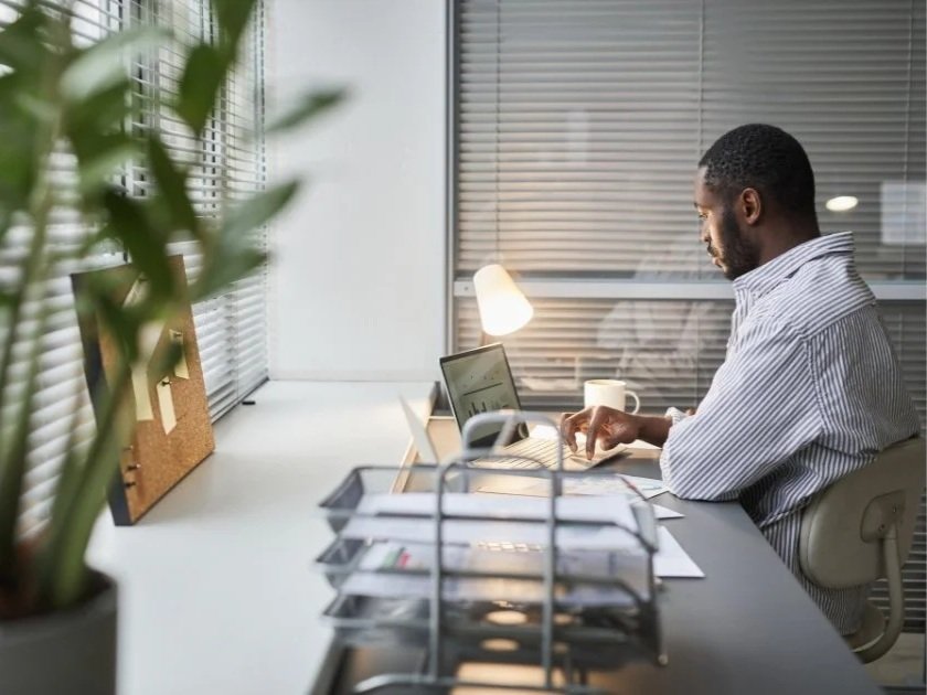 A man working at a desk with a laptop, a desk lamp, a coffee mug, and office supplies in a well-lit room with blinds.