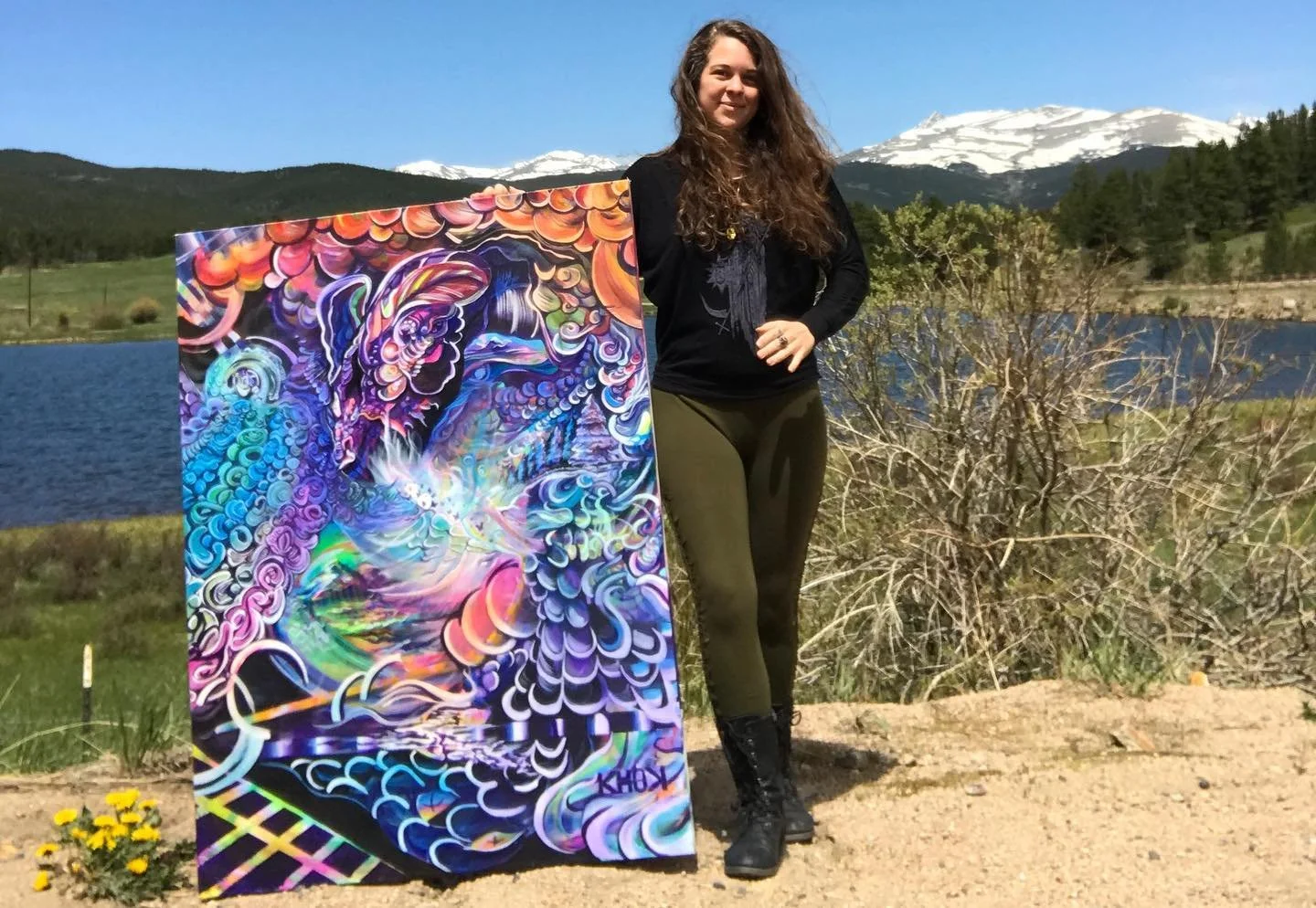 A woman with long curly hair standing outdoors next to a colorful abstract painting, with a lake, mountains, and snow-capped peaks in the background.
