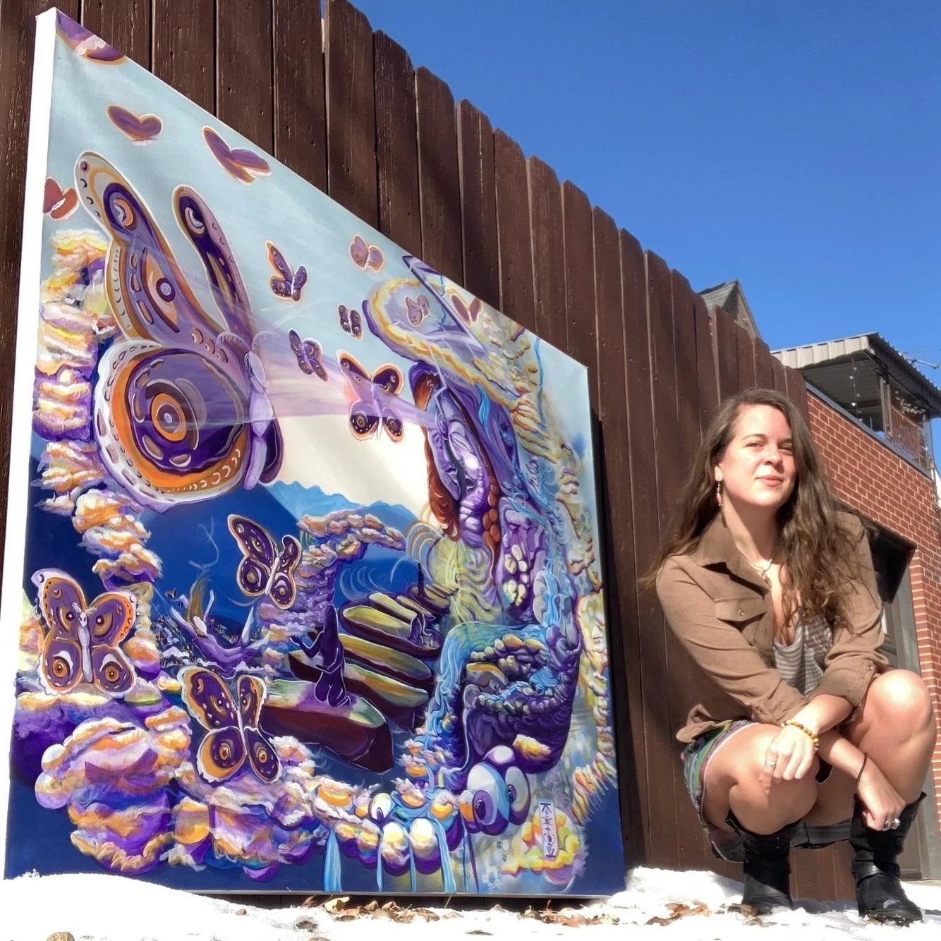 A woman with long, wavy brown hair sitting next to a colorful butterfly-themed painting outdoors against a wooden fence and a brick building. The sky is clear and blue.