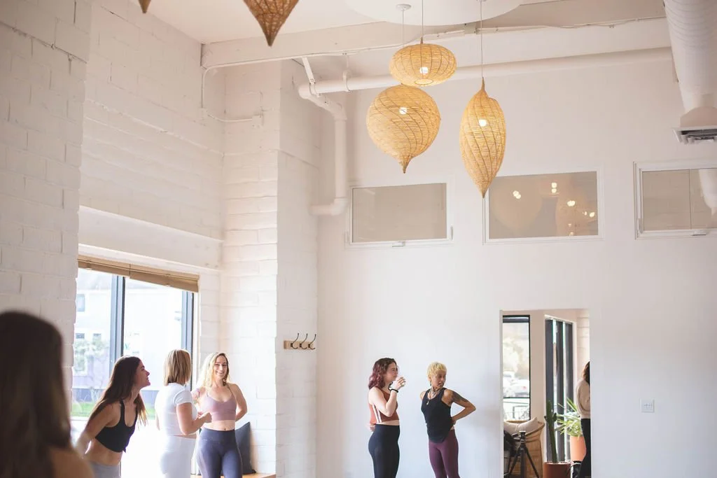 Group of women in workout clothes participating in a fitness class in a bright, modern studio with hanging wicker lights and large windows.