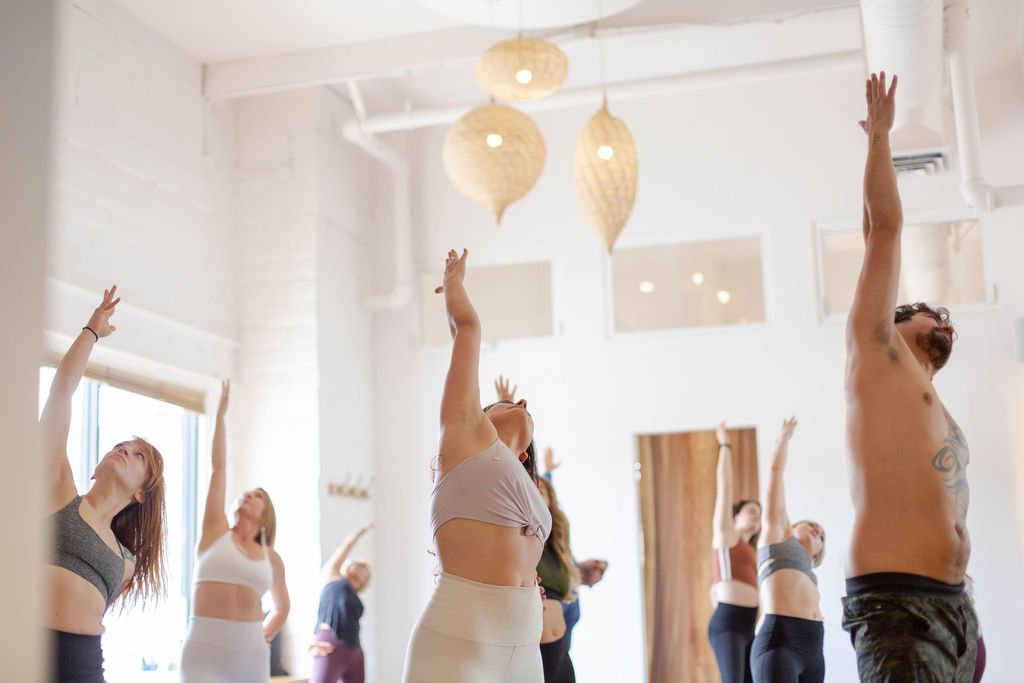 Group of people holding yoga pose in a well-lit studio with hanging paper lanterns.