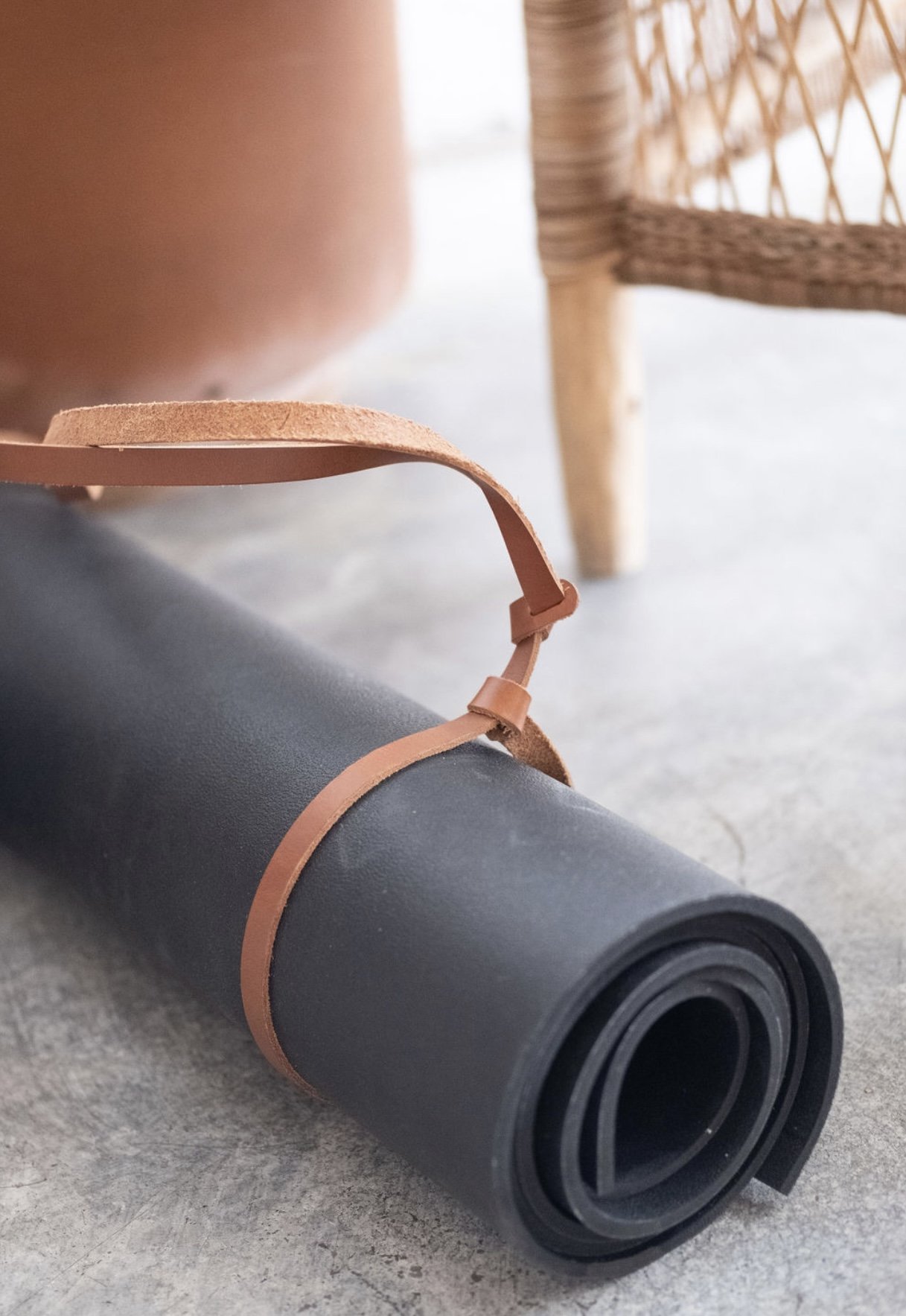 A yoga mat with a leather strap lying on a concrete floor, with a wicker chair and a large clay pot in the background.