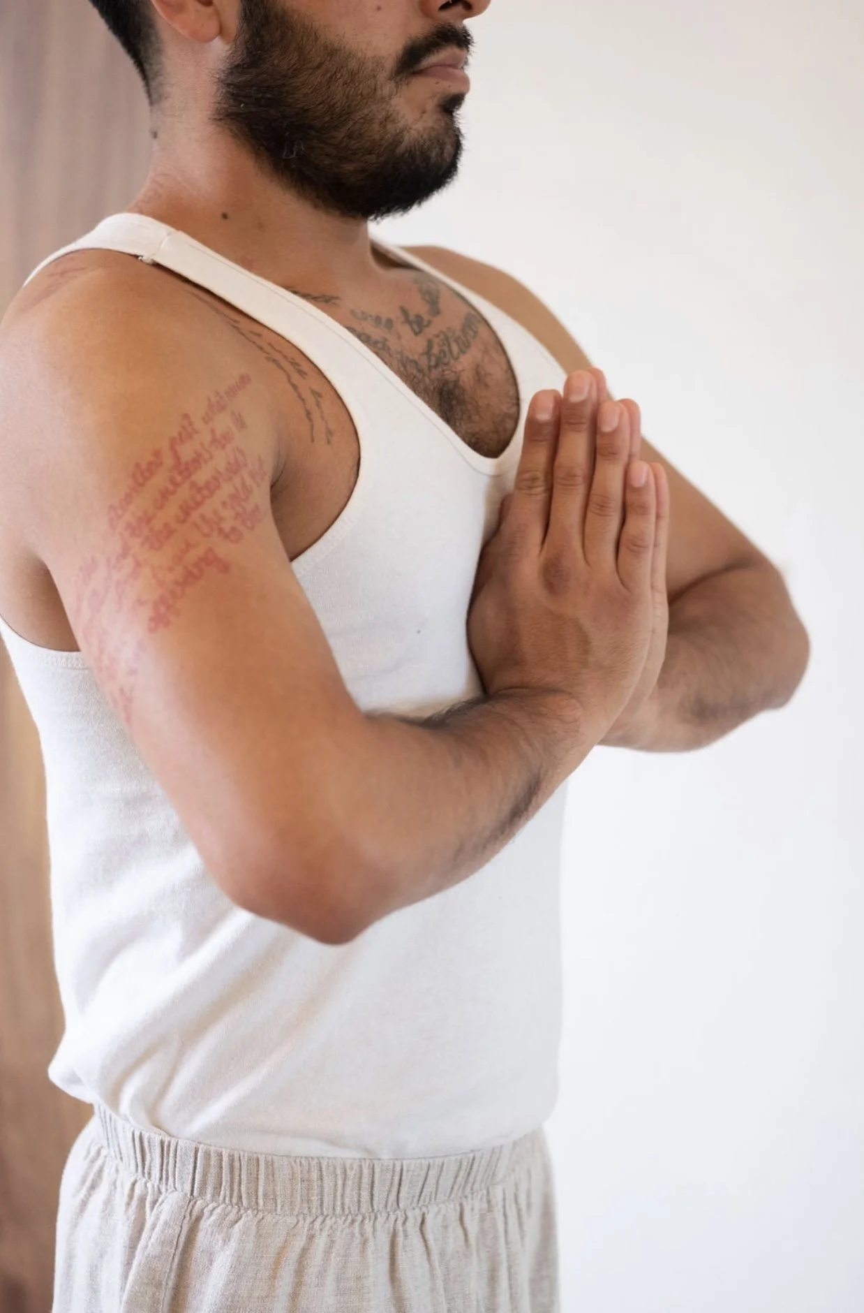 A man doing yoga with eyes closed, hands pressed together in prayer position, shirtless with tattoos, wearing beige pants, standing against a plain wall.