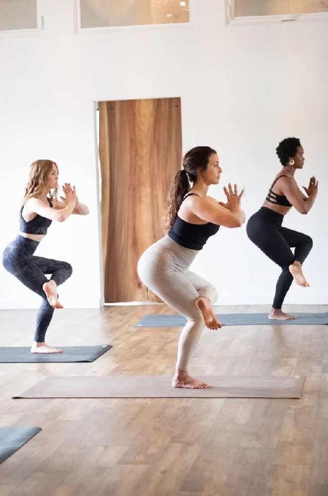 Three women practicing yoga indoors, standing on mats and balancing with knees bent and hands raised in a prayer position.