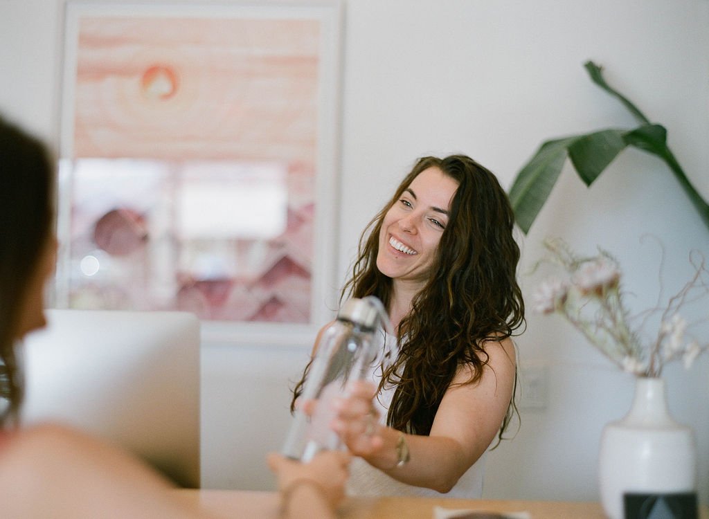 A woman with long, curly brown hair smiling and holding a hairdryer, sitting at a table in a bright room with artwork and a large plant in the background.