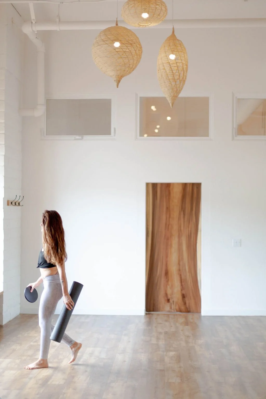 A woman holding a yoga mat and foam roller walking in a minimalist, brightly lit room with white walls and wooden flooring, hanging wicker pendant lights, and a wooden door.