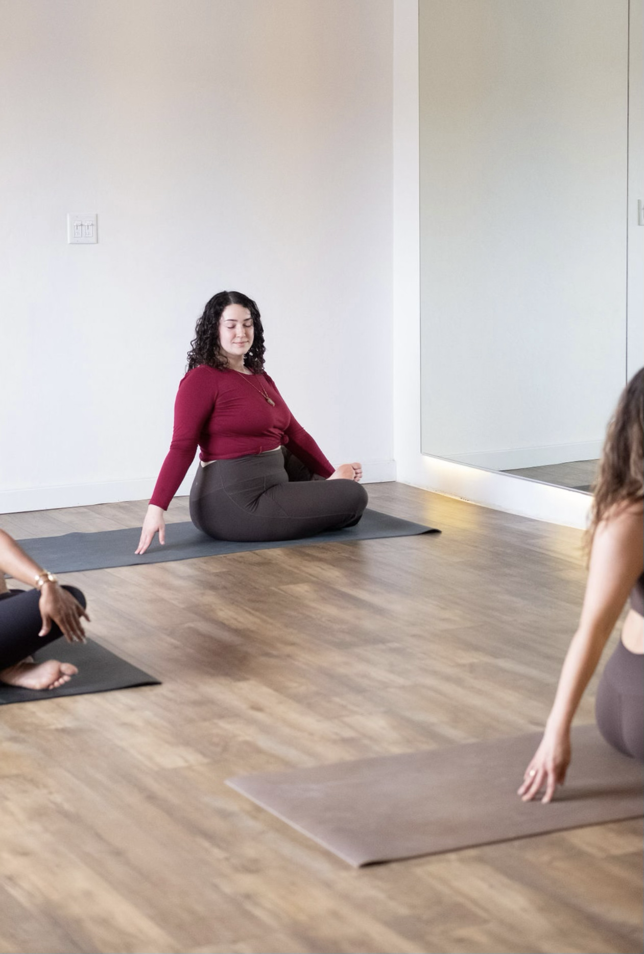 Women participating in a yoga class, sitting on mats, with one instructor in a red top leading a pose in front of a mirror.