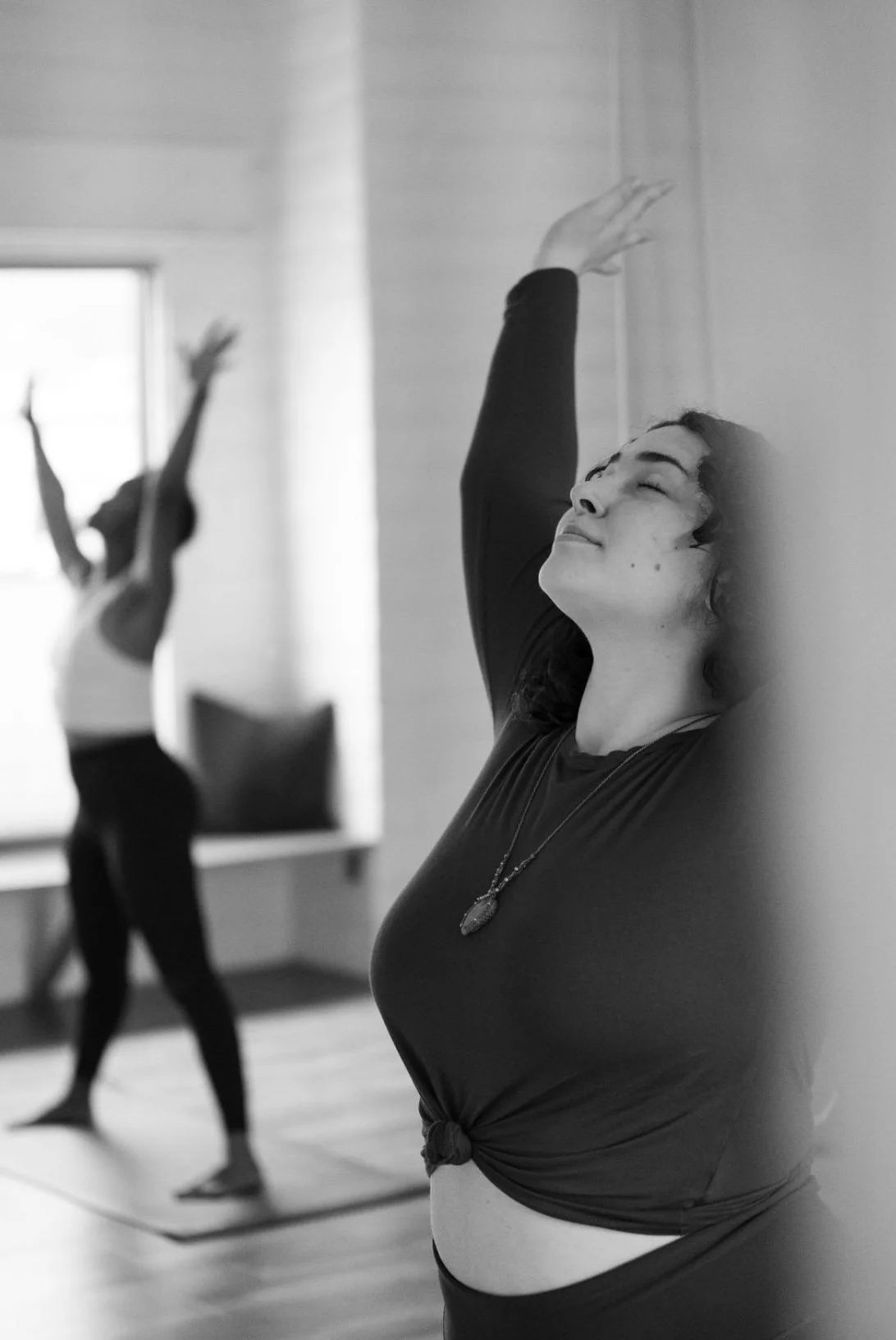 Two women practicing yoga, one in the foreground with eyes closed and arm raised, the other in the background in a similar pose, in a well-lit room.