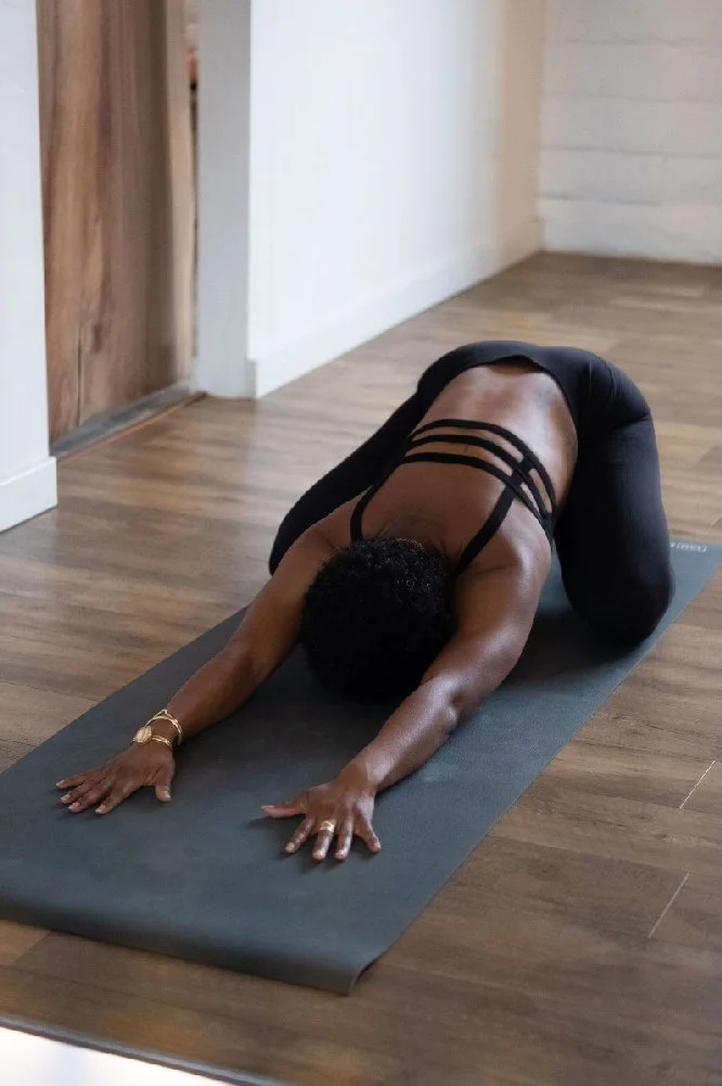 A person practicing yoga on a black mat, positioned in child's pose with knees bent, arms extended forward, and head down.