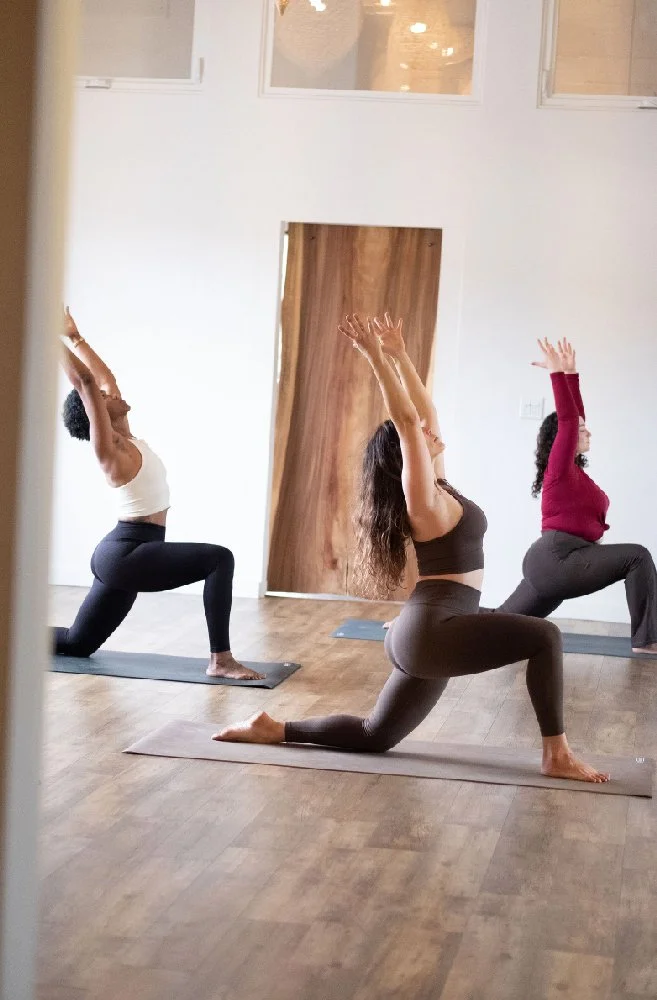 Three women practicing yoga in a bright, minimalistic room with hardwood floors and large windows, performing a low lunge pose with arms raised.