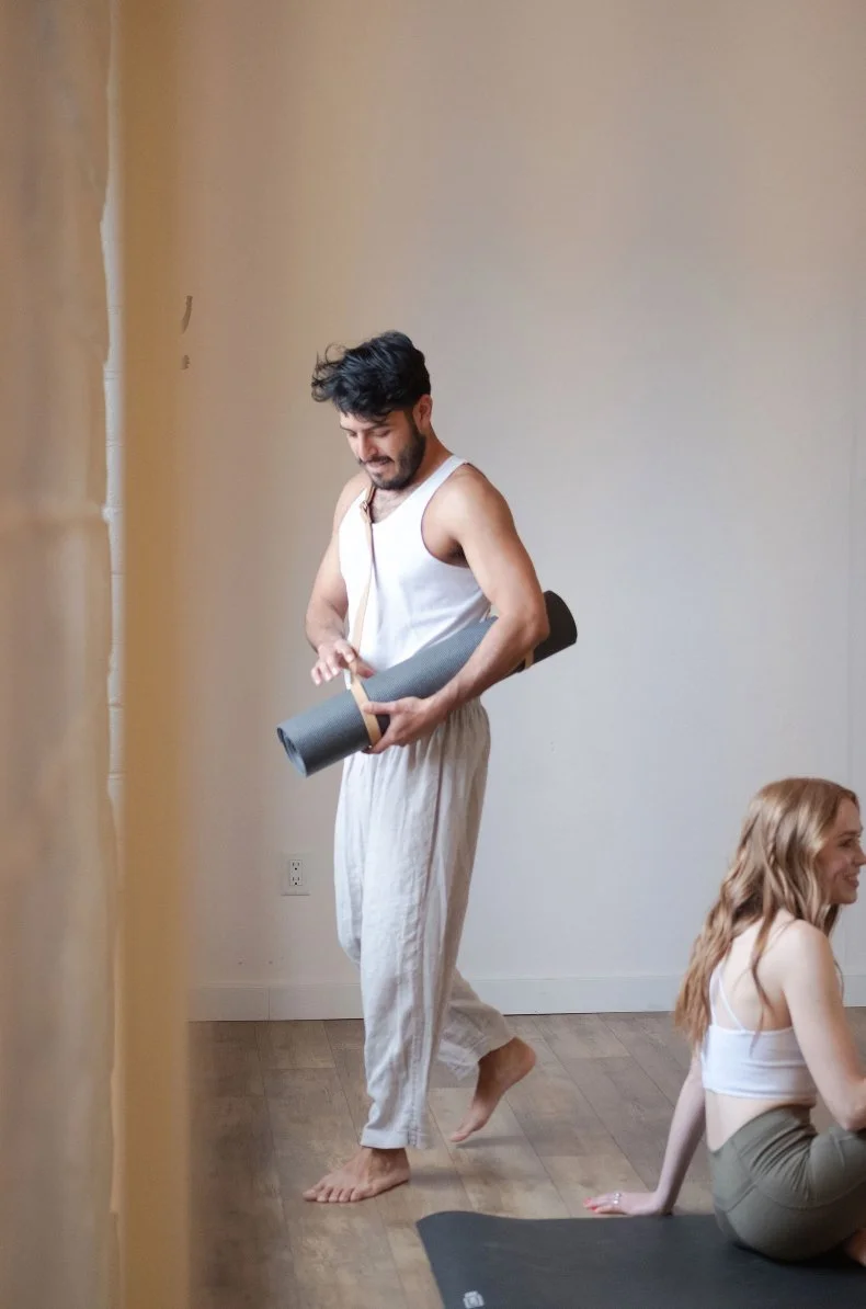 A man standing in a room holding a yoga mat with a woman sitting on a yoga mat nearby.