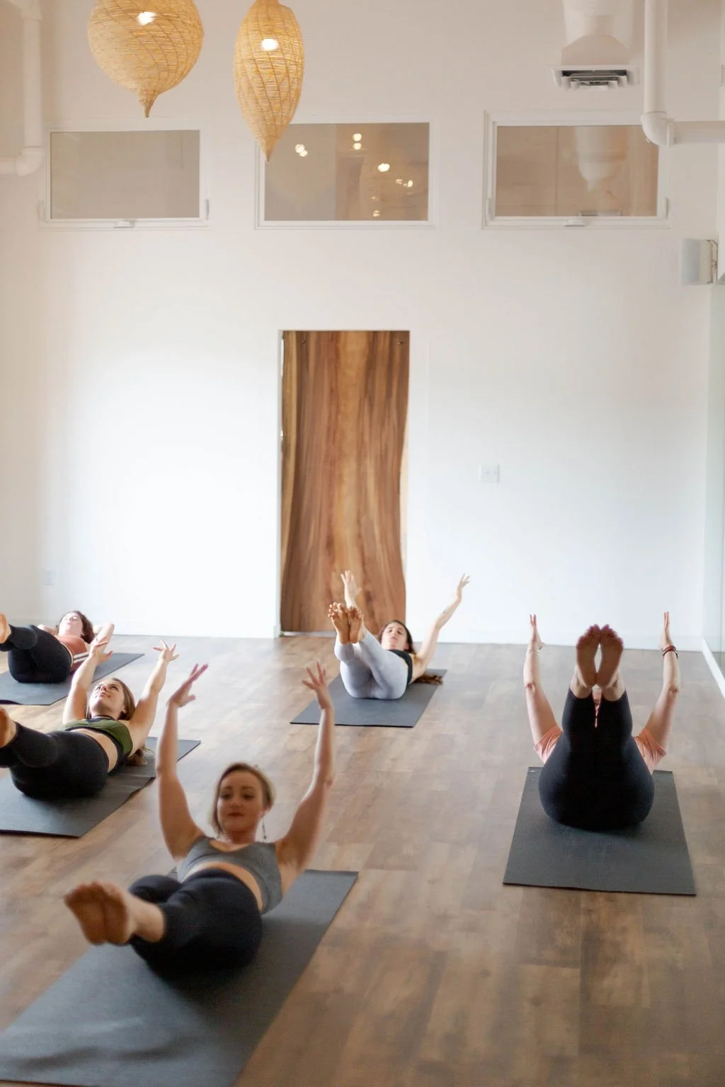 Group of women practicing yoga on mats in a bright, modern studio with white walls, wooden flooring, and decorative hanging light fixtures.