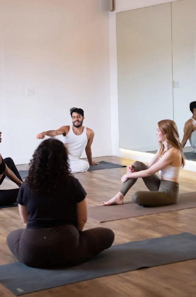 A diverse group of people participating in a yoga class in a studio with wooden floors and a large mirror, seated on yoga mats.