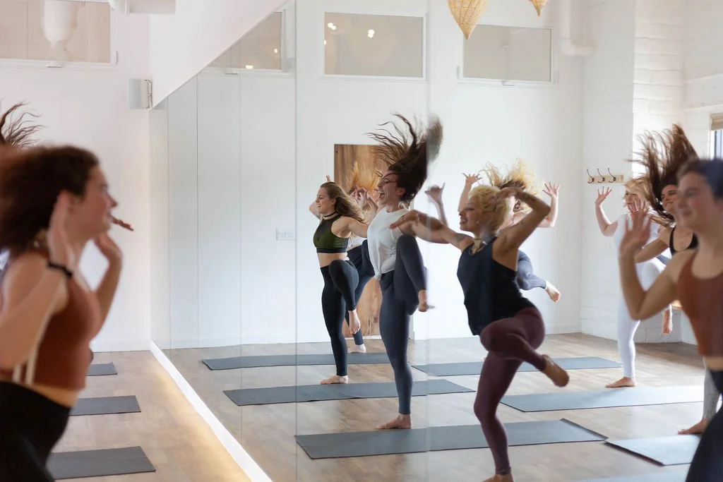 Group of women participating in a dance or yoga class, jumping with hair flying, in a bright studio with a mirrored wall.