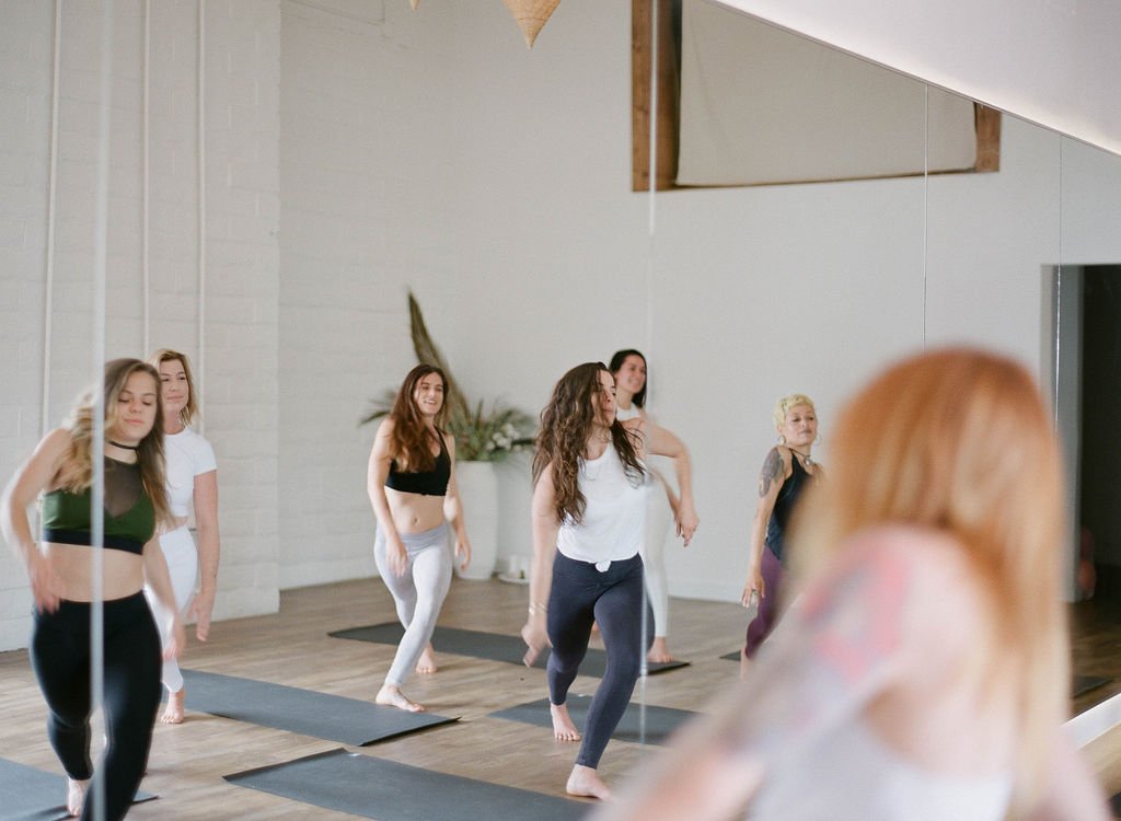 Group of women participating in a yoga class in a studio with wooden floors and white walls, some are standing on yoga mats, and a large mirror reflects their images.