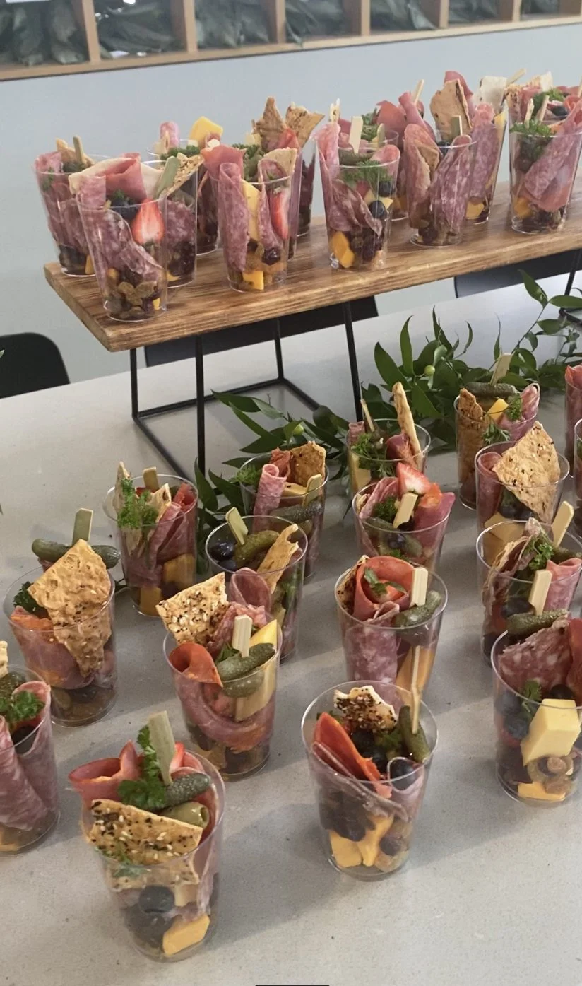 Individual appetizer cups filled with cured meats, cheeses, crackers, and garnishes, arranged on a table with a wooden stand and greenery.
