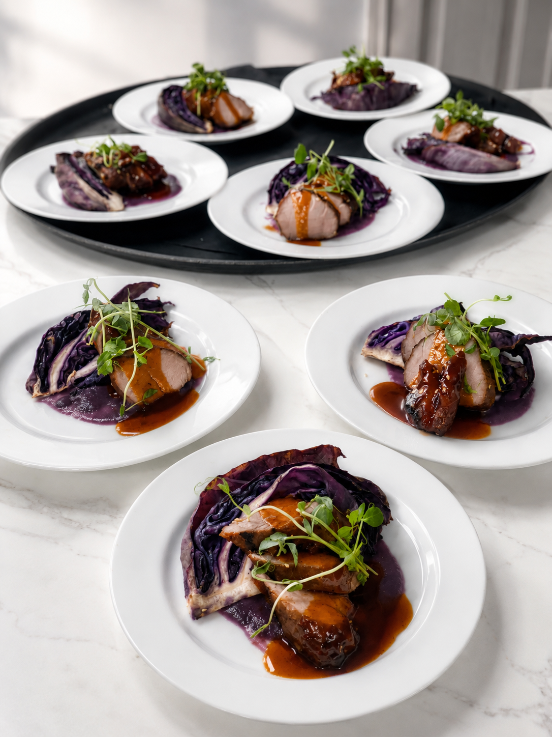 Six plates of sliced pork tenderloin with purple cabbage and microgreens, drizzled with brown sauce, served on white plates on a marble surface and a black tray.