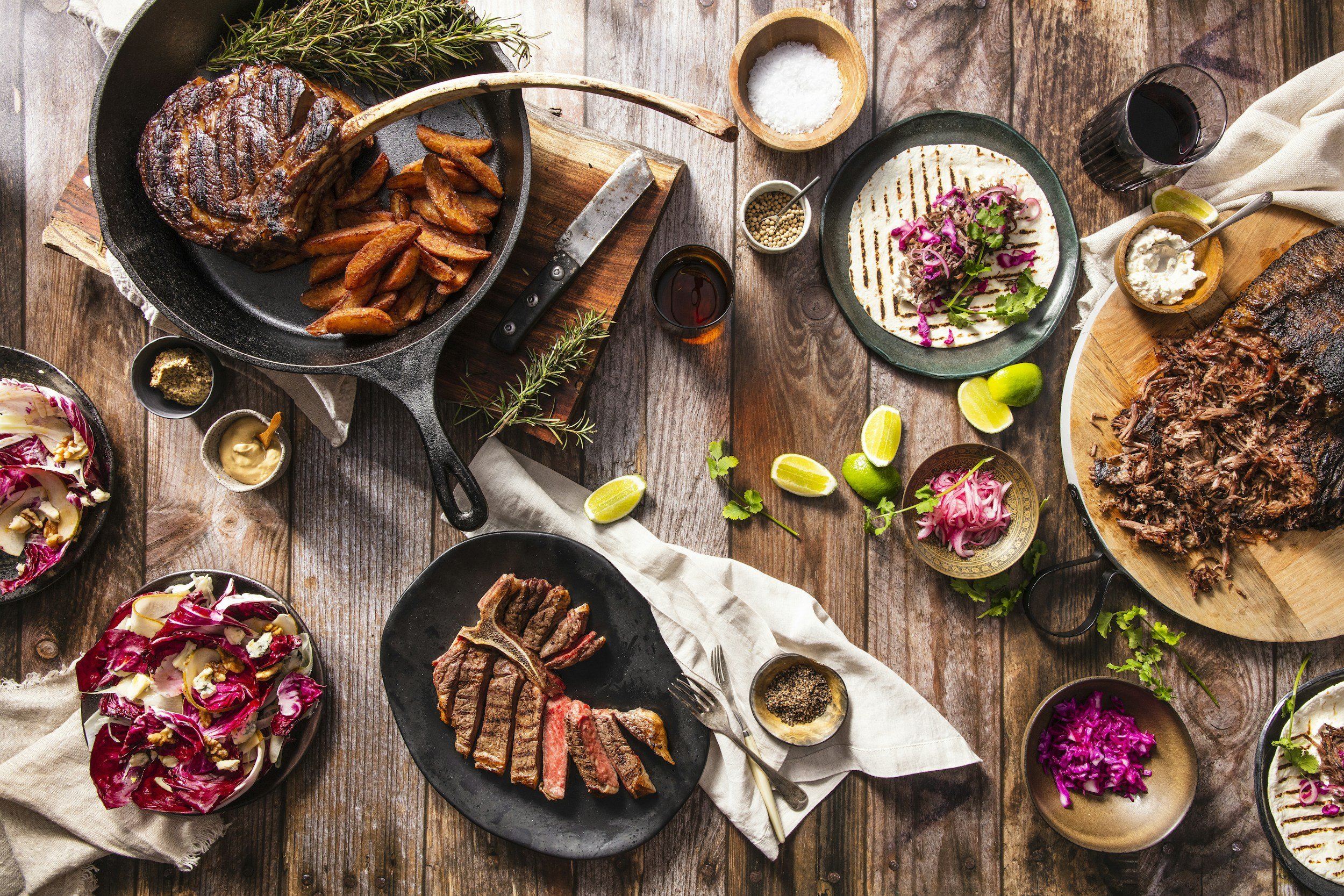 A rustic wooden table with various dishes including grilled steak, roasted potatoes, grilled vegetables, salad, and sauces, accompanied by lime wedges and glasses of red wine.