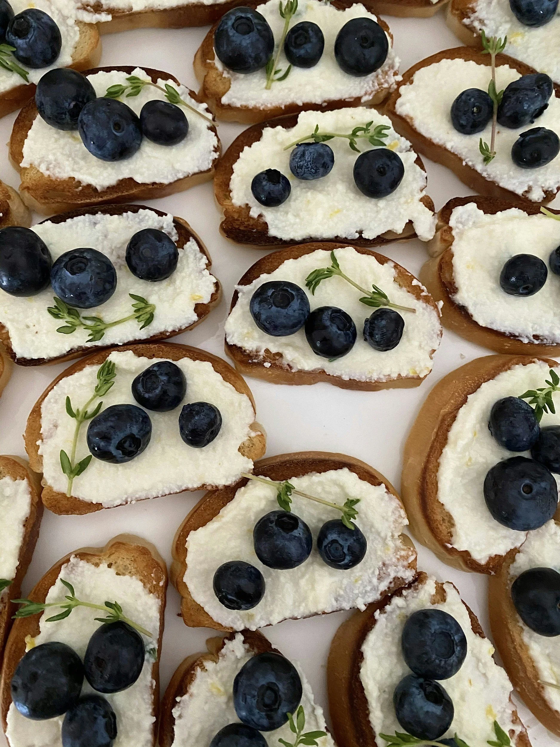 Open-faced bread slices topped with cream cheese, blueberries, and small green herbs.