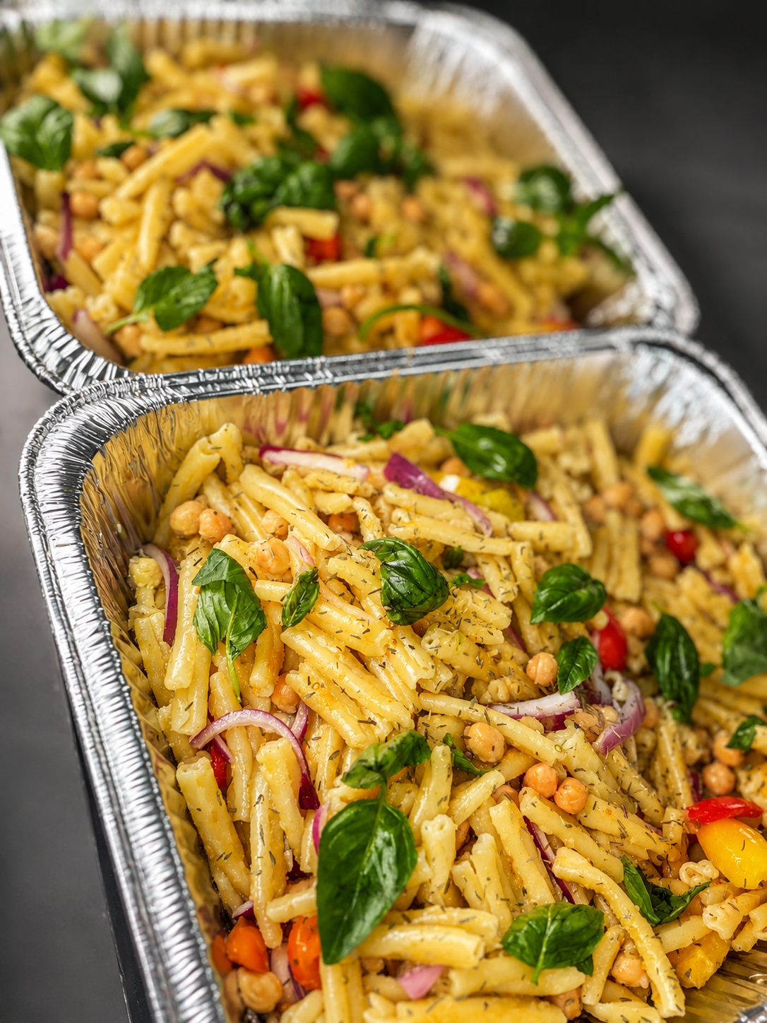 Two aluminum foil containers filled with pasta salad, garnished with fresh basil leaves, chopped red onions, and chickpeas.