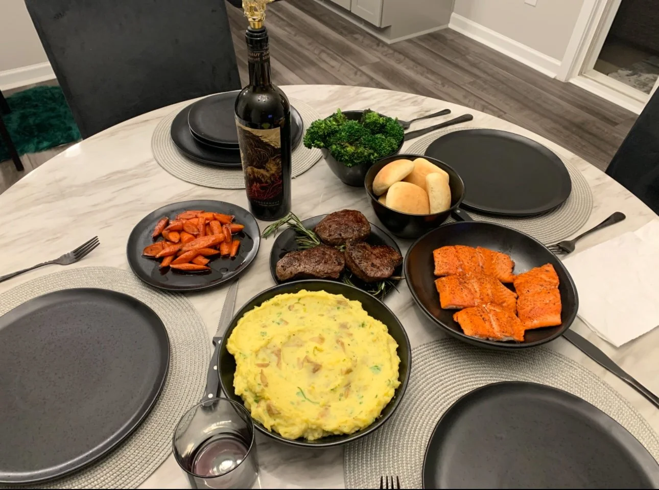 A dinner table set with black plates, a bowl of mashed potatoes, cooked salmon, grilled beef patties, roasted carrots, broccoli, a bottle of red wine, and a glass of wine.