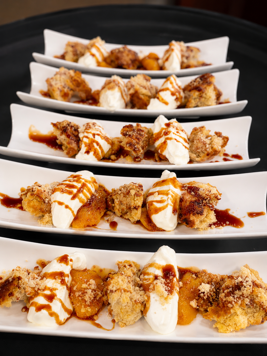 Four white rectangular plates with dessert pieces topped with whipped cream and caramel sauce, arranged on a black tray.