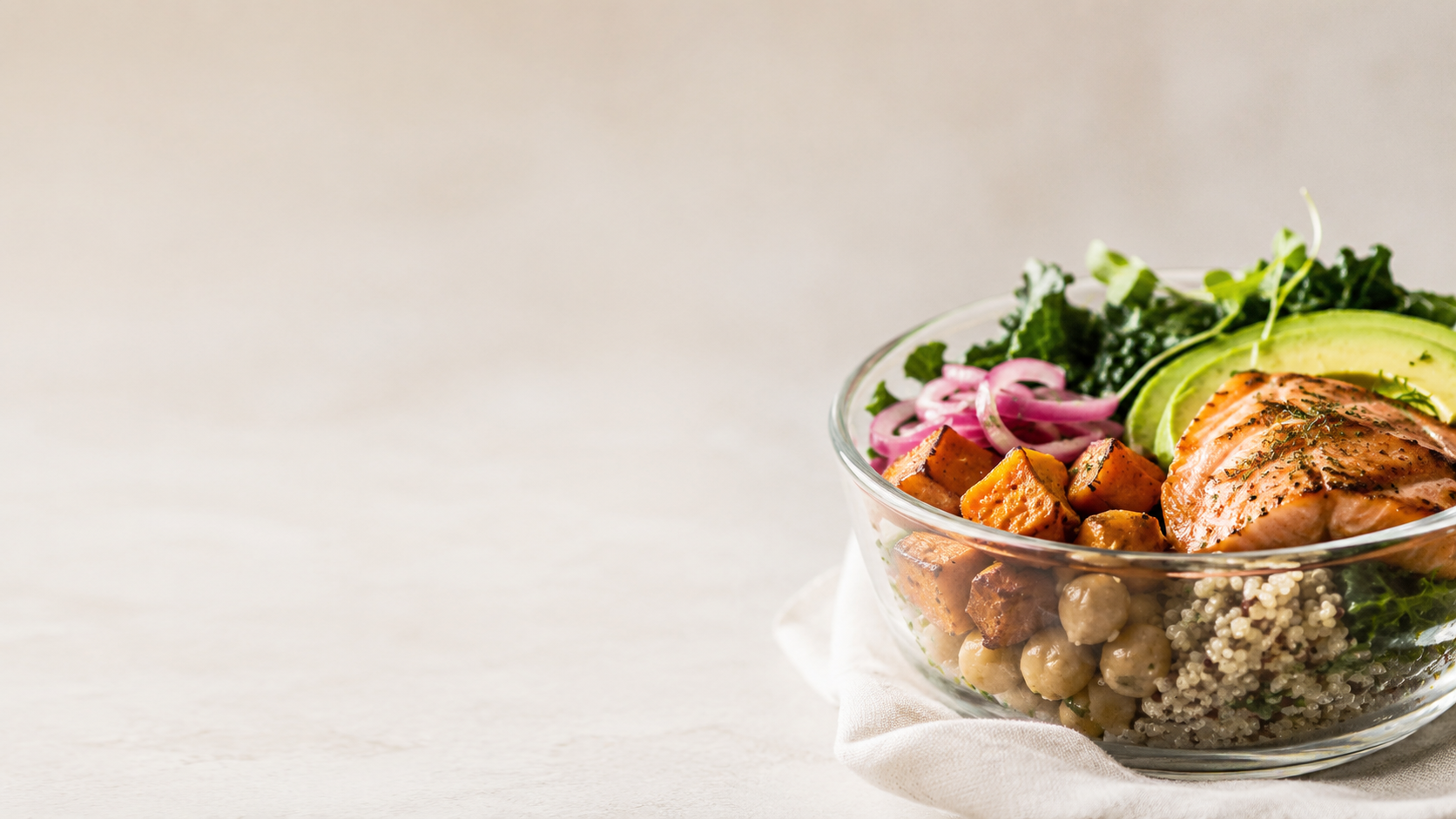A glass bowl of colorful salad with greens, grilled chicken, chickpeas, curry tofu, quinoa, and pickled onions on a white cloth.