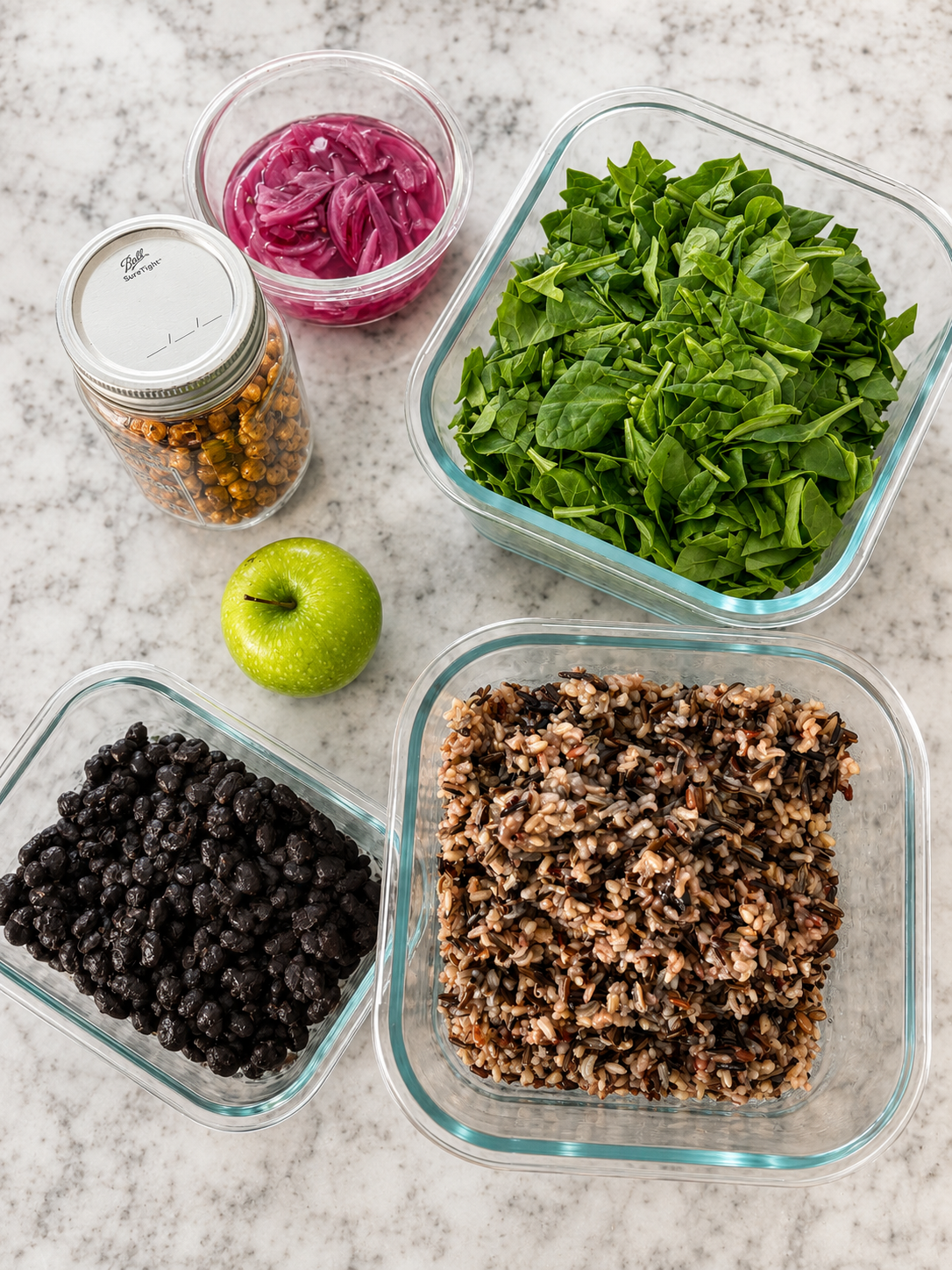 A green apple, cooked black beans, wild rice, kale, pickled red onions, and pickled okra arranged in glass containers on a white countertop.