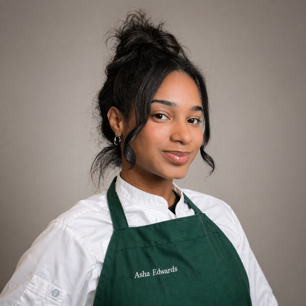 A woman with dark, curly hair in a messy updo, wearing a white chef's coat and a green apron with her name, Asha Edwards, embroidered on it. She is smiling softly against a plain gray background.