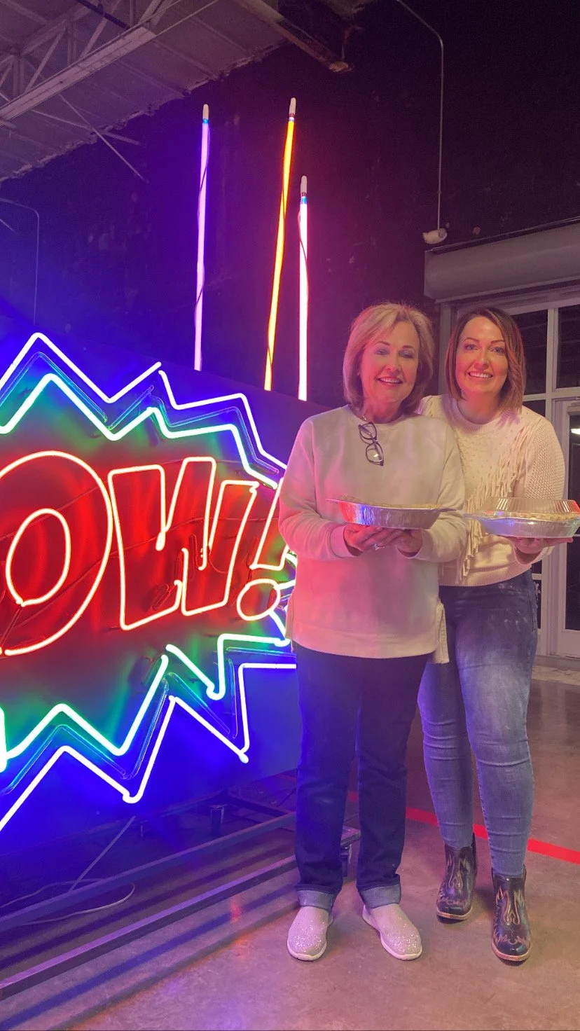 Two women smiling and holding food containers, standing in front of a neon sign with colorful lights and the word 'POW!' in red, with neon lightning bolts above.