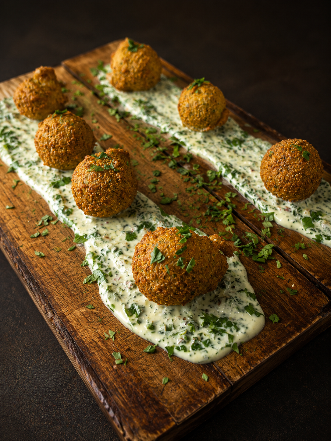 Six falafel balls on a wooden platter, served over a creamy herb sauce with chopped herbs and garnished with chopped parsley.
