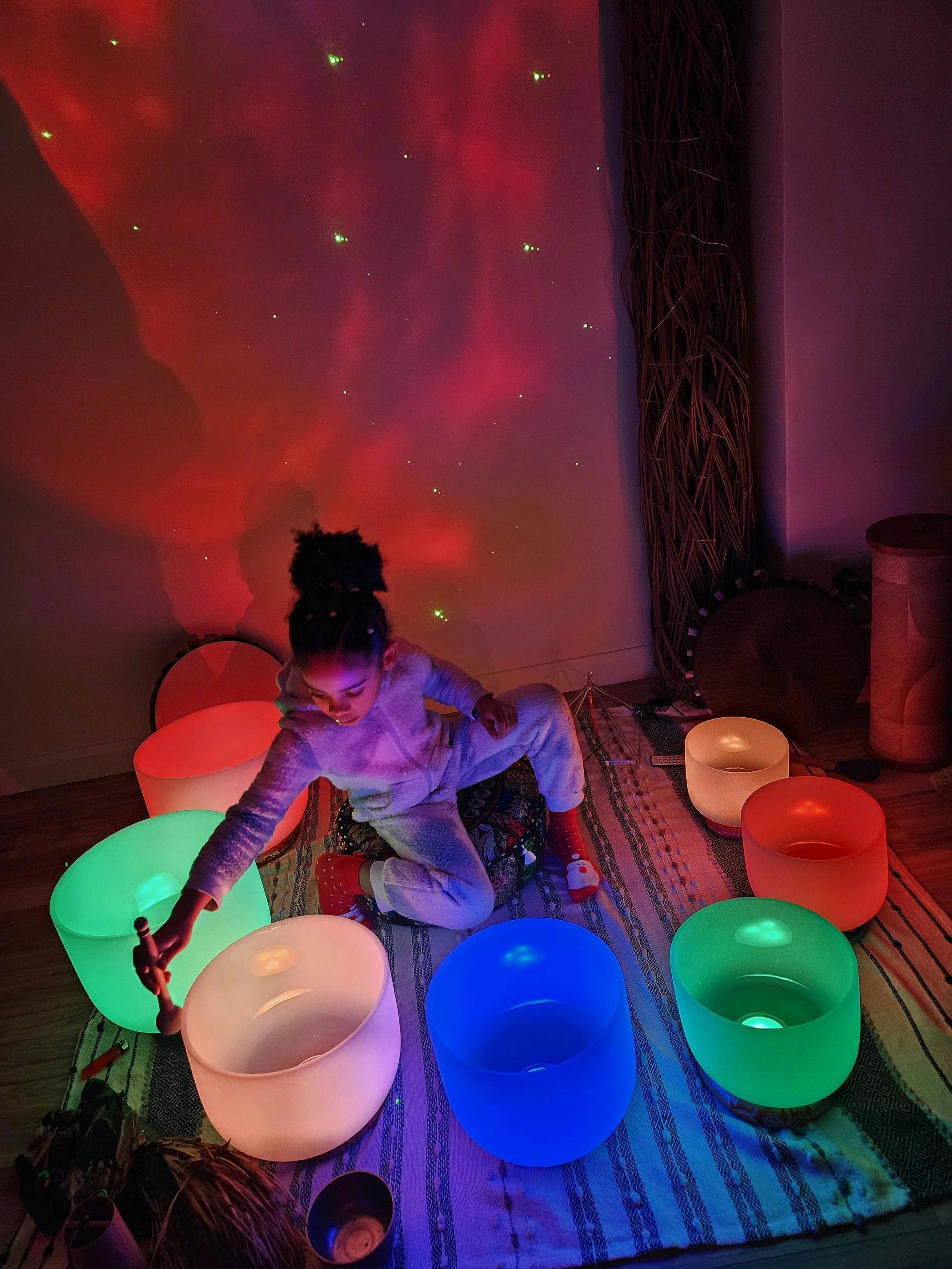 A young girl is sitting on a colorful, striped rug in a dimly lit room with glowing, multicolored bowls around her. She is reaching into one of the bowls, which are illuminated in vibrant green, pink, blue, and orange colors. The room has a starry sky and galaxy projection on the wall and some dark curtains and furniture in the background.
