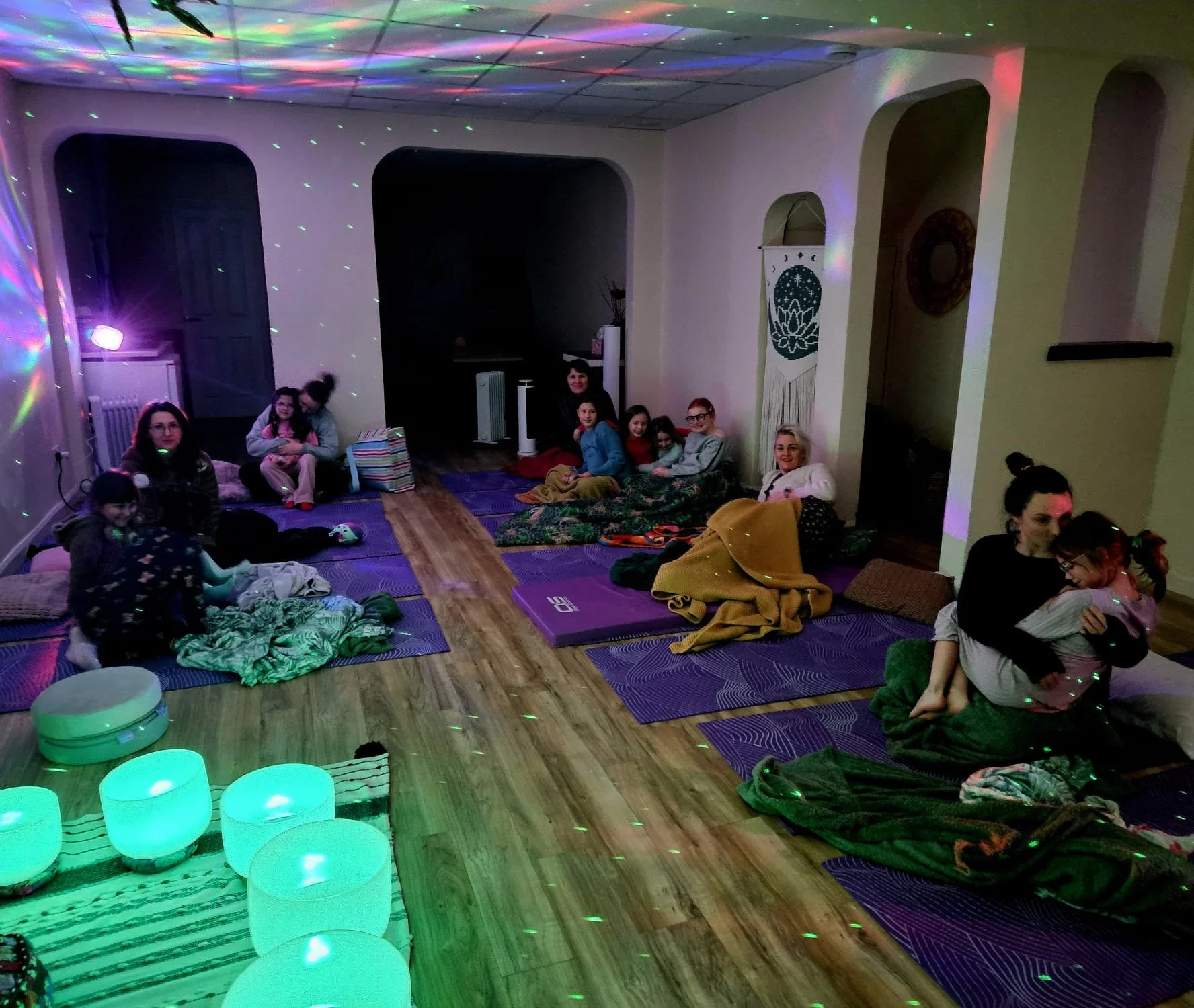 A group of children and adults sitting on mats and blankets in a room with colorful disco lights, enjoying a relaxed gathering.
