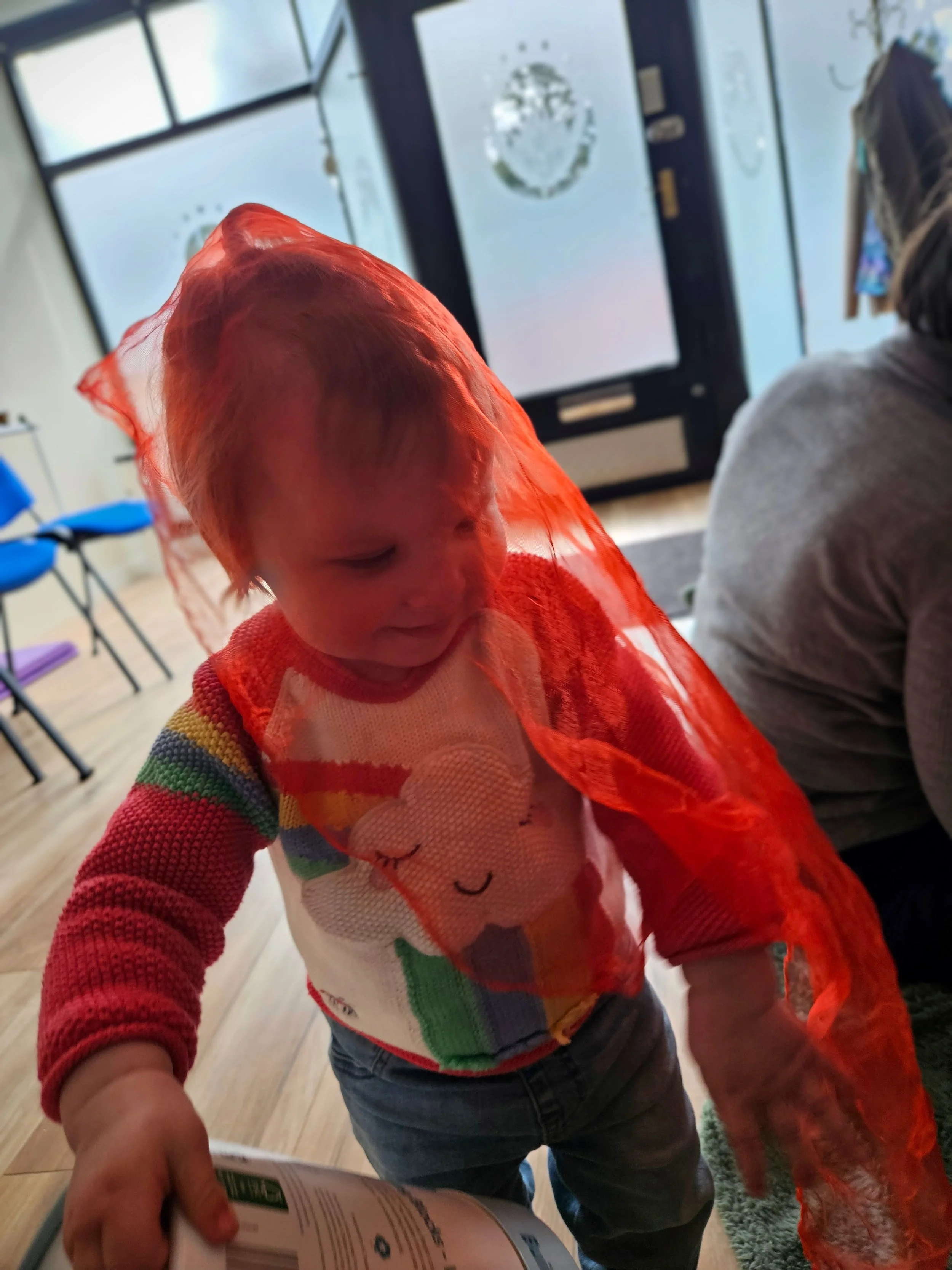 A young child with light skin, wearing a colorful sweater with a smiling face design, is looking down at a brochure or booklet. The child has a red sheer fabric draped over their head. In the background, there is a glass door with a nature scene reflected on it, and a person with long hair and a hat sitting nearby.
