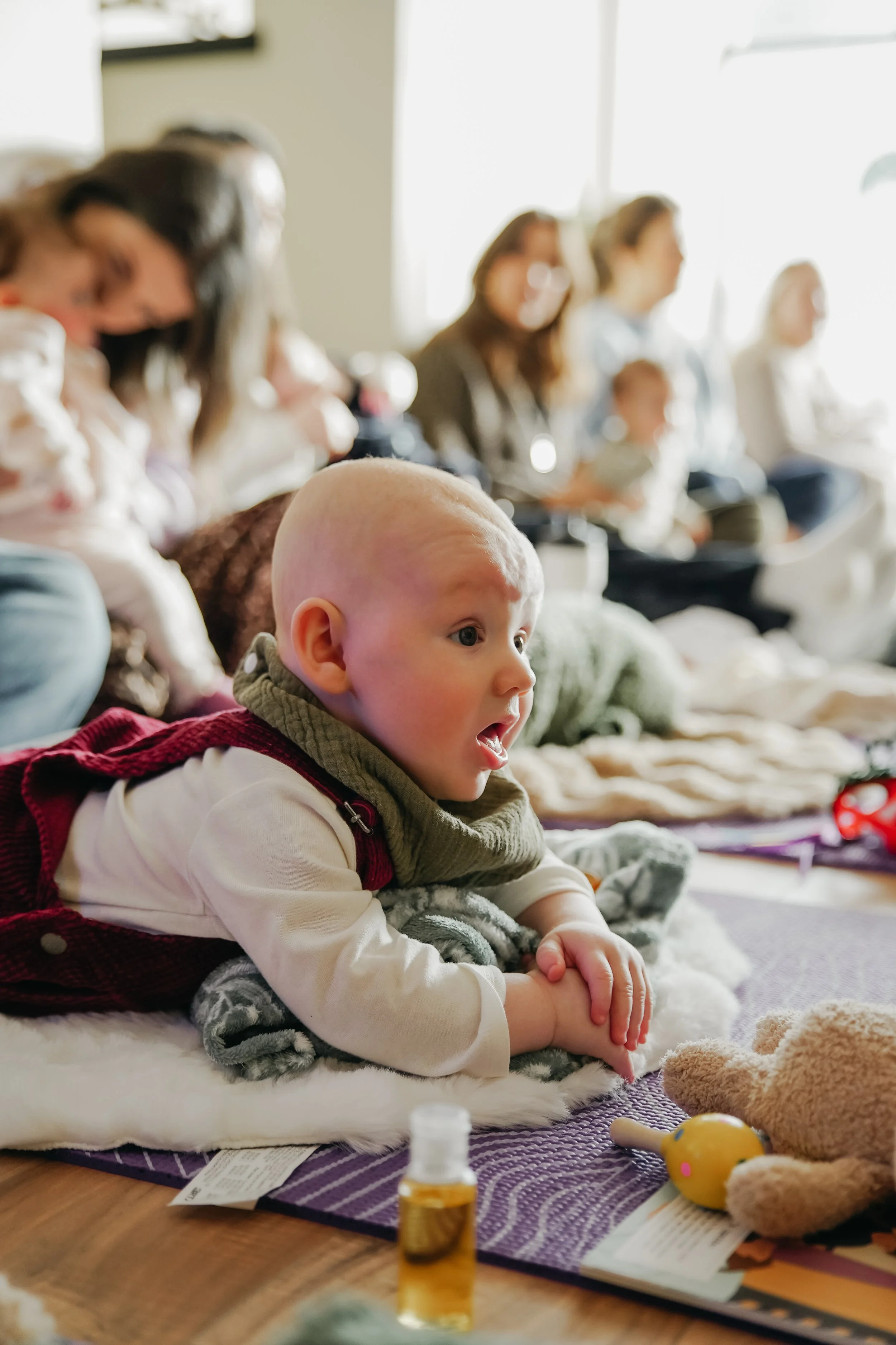 A young child with a bald head and light skin lying on a blanket, watching attentively with an open mouth, amongst toys, in a well-lit room with a group of adults and children in the background.