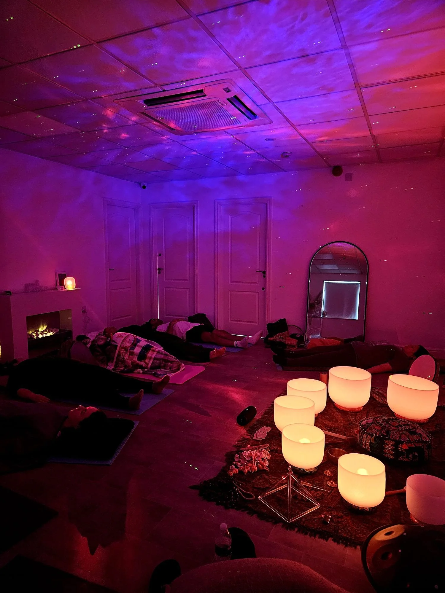 A dimly lit room with purple and red lighting effects. There are several people lying on mats on the floor, possibly participating in a relaxation or meditation session. The ceiling has a purple hue with small star-like reflections. There is a mirror on the right side of the room, and large white bowls with candles on a rug in the foreground. The atmosphere appears calm and peaceful.