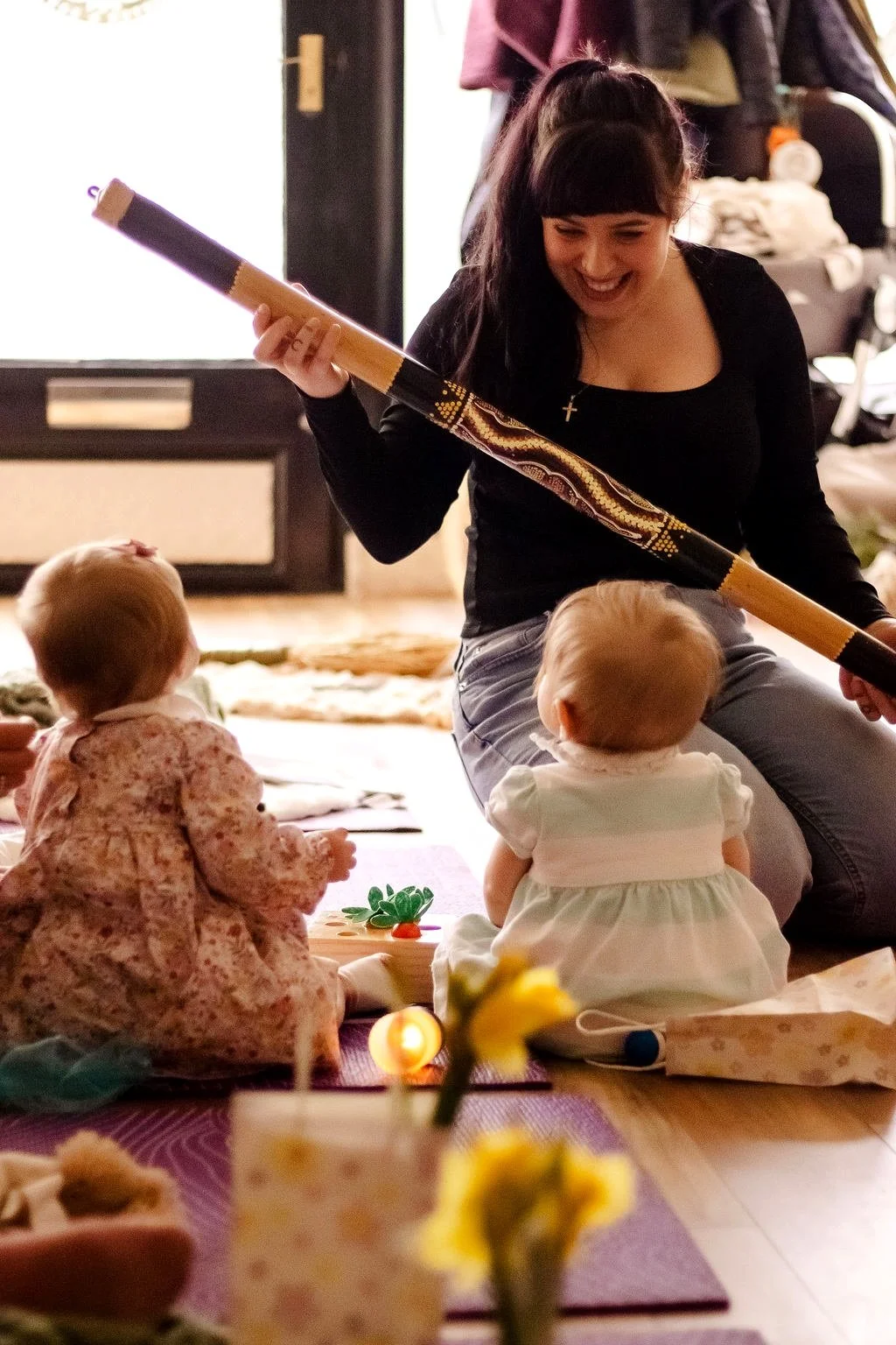A woman sits on the floor with two young children, one girl and one baby, during a lively indoor gathering. The woman is holding a long decorated stick and smiling warmly at the children. The scene is warm and festive with soft indoor lighting.
