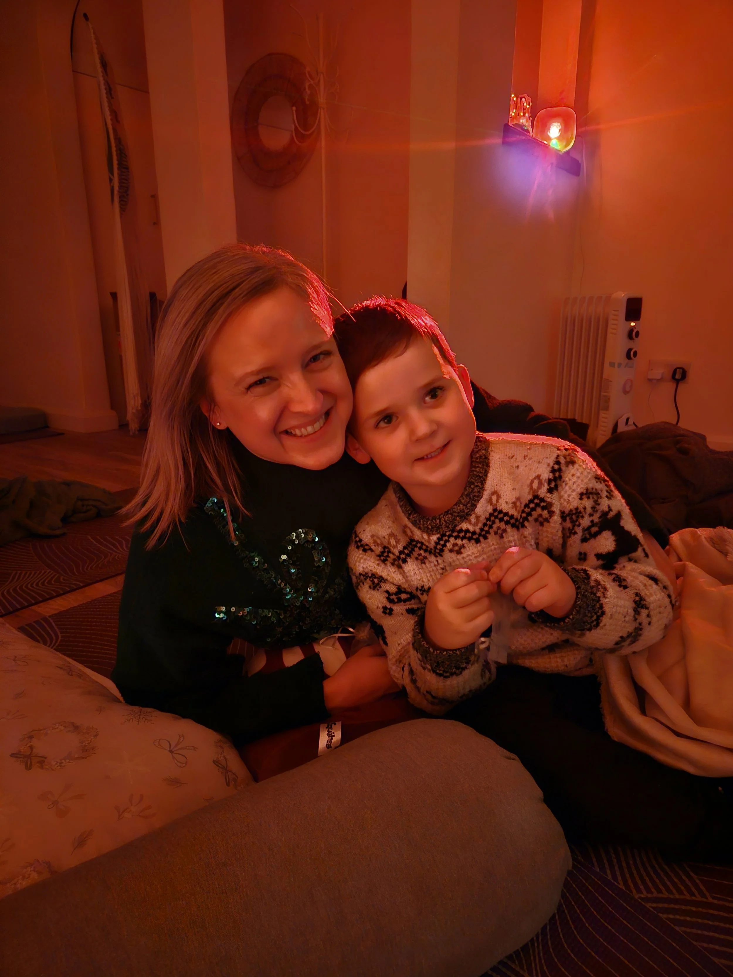 A woman and a young boy smiling and hugging on a couch in a dimly lit room with colorful lighting. The woman has blonde hair and the boy has short brown hair, wearing a patterned sweater. There are cushions on the couch and a heater on the wall in the background.
