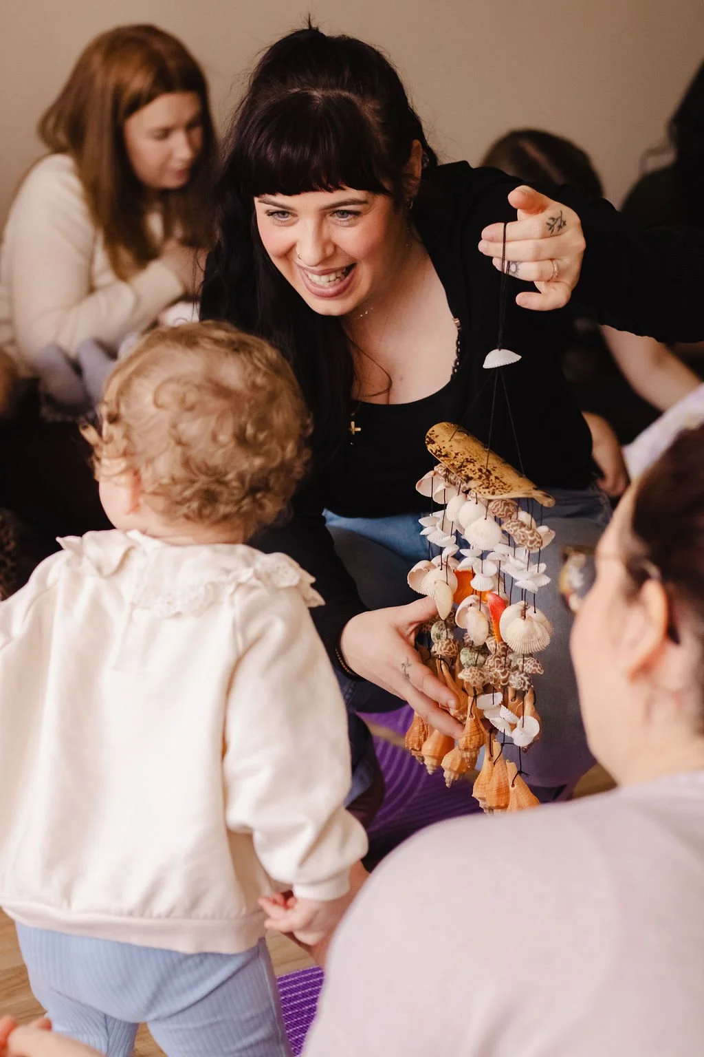 A woman with dark hair and a black top is smiling and showing a mobile hanging decoration to a young child with curly blonde hair wearing a light-colored jacket. Other people are visible in the background.