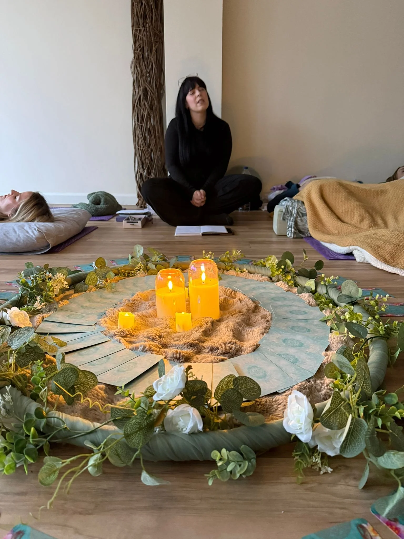 A woman with black hair and black clothing kneels on the floor with her eyes closed and hands clasped. She is in front of a table with candles and greenery arranged in a floral centerpiece. There are two people lying on the floor on either side of her, covered with blankets.