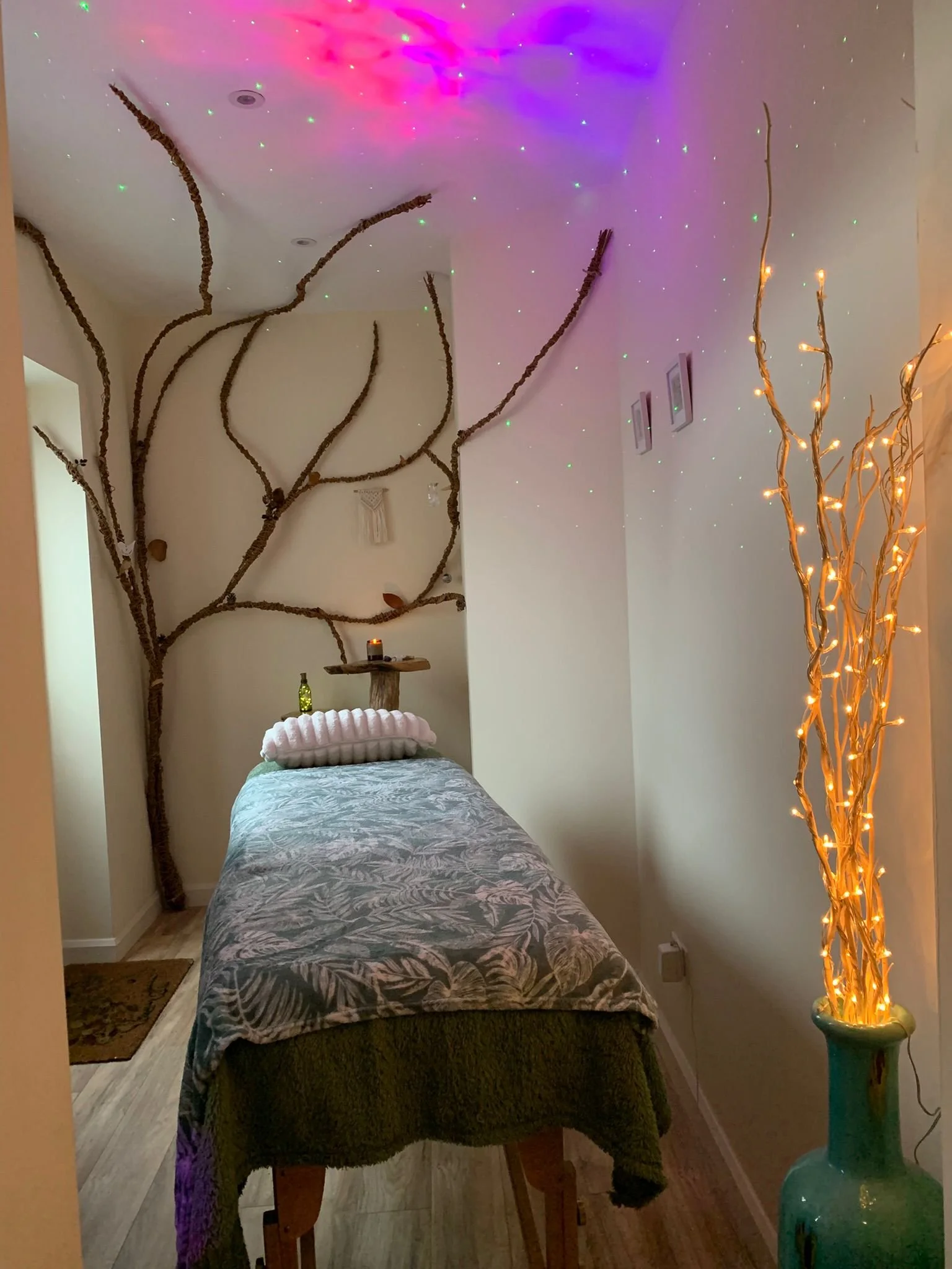 A cozy bedroom featuring a bed with a gray and white animal print blanket, decorative pillows, and a small green bottle on the bedside table. The room has artistic branch decor on the wall, star-like lights on the ceiling, and a tall vase with glowing, twisted branches wrapped in small lights to the right.