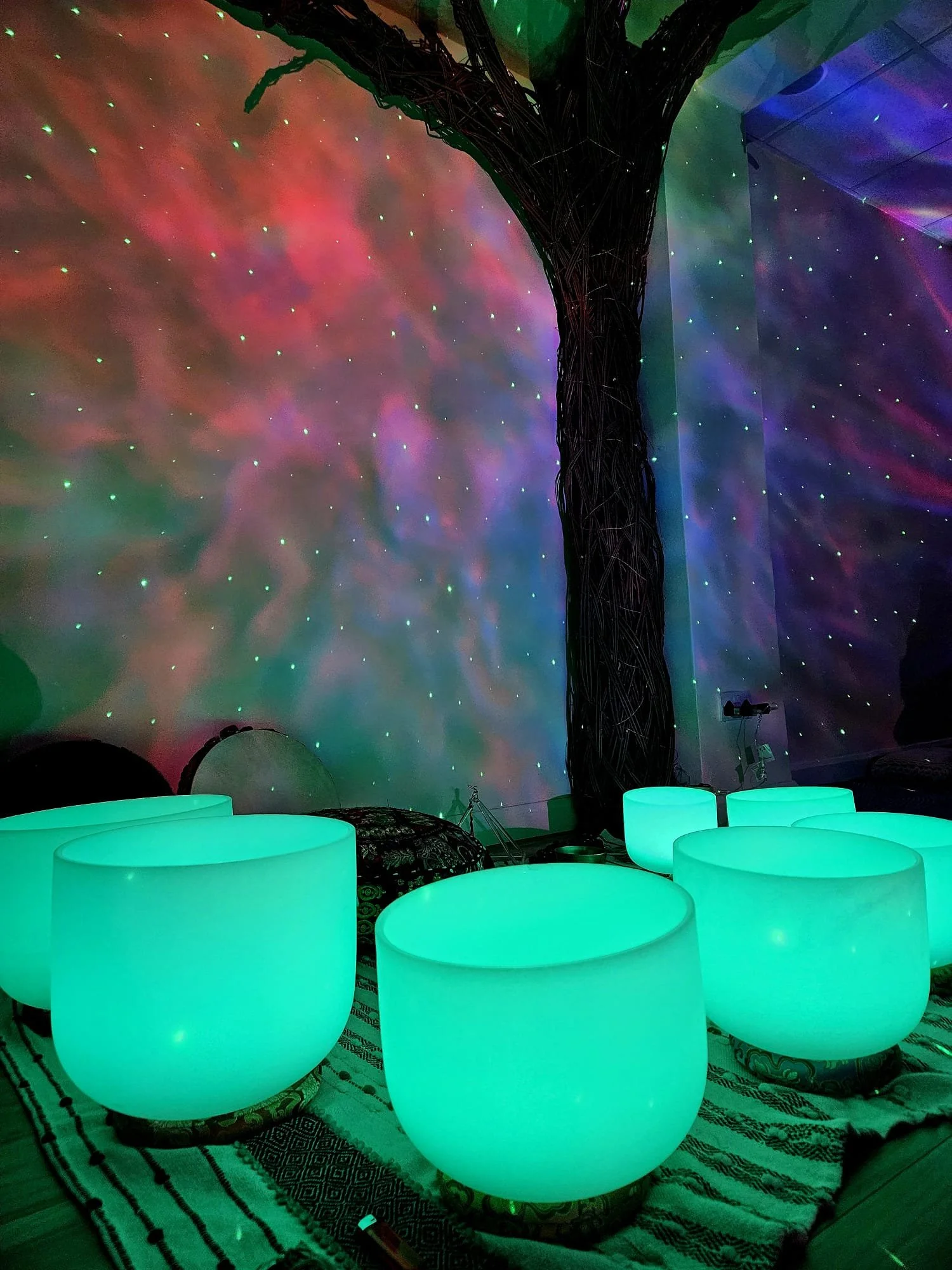 A room with glowing teal-colored spherical seats, a large illuminated artificial tree, and a wall projection of colorful, star-like lights and nebula patterns.