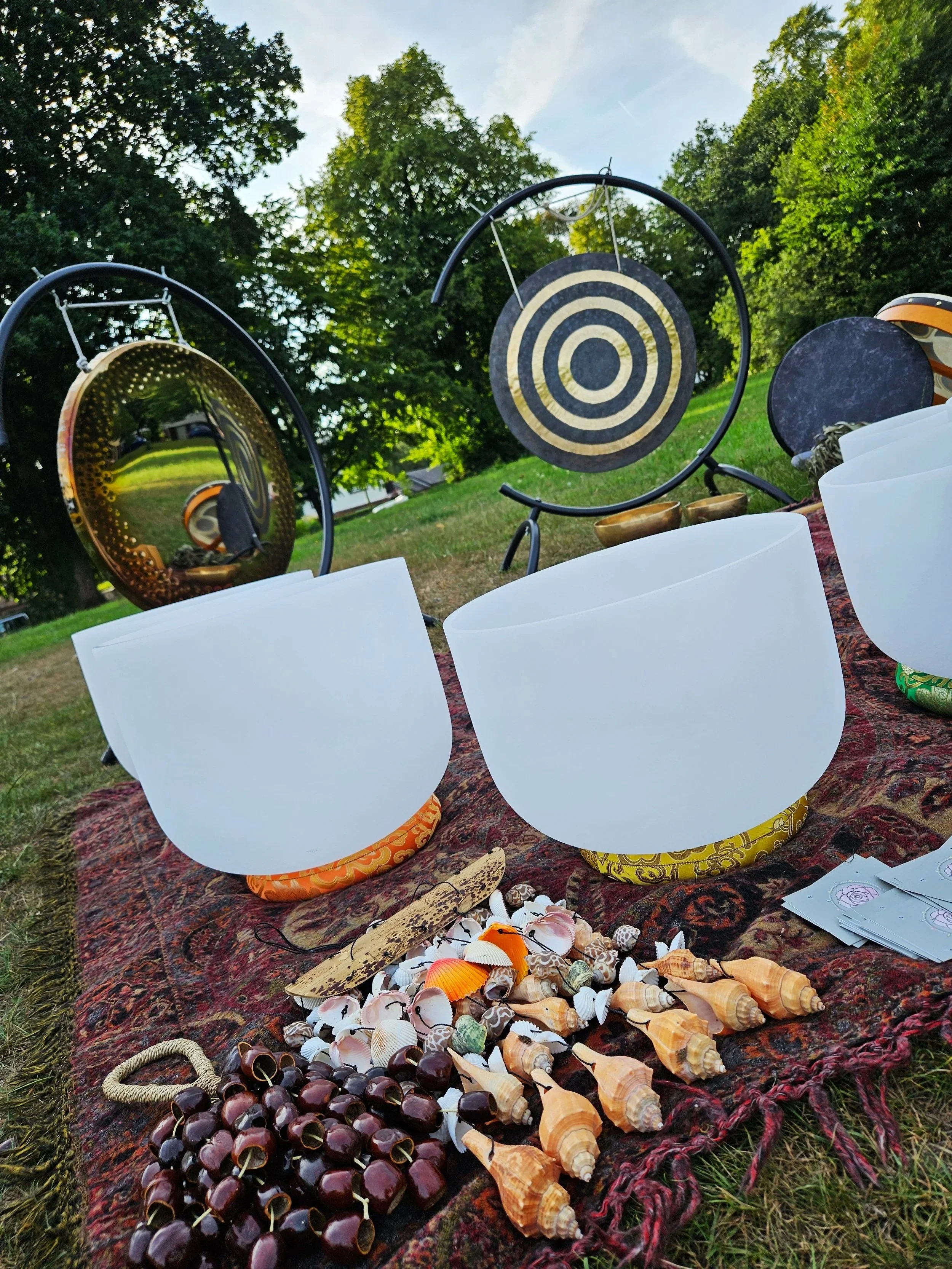A collection of seashells, coconuts, and other objects on a patterned cloth spread over grass, with gongs hanging in the background, outdoors with trees and blue sky.