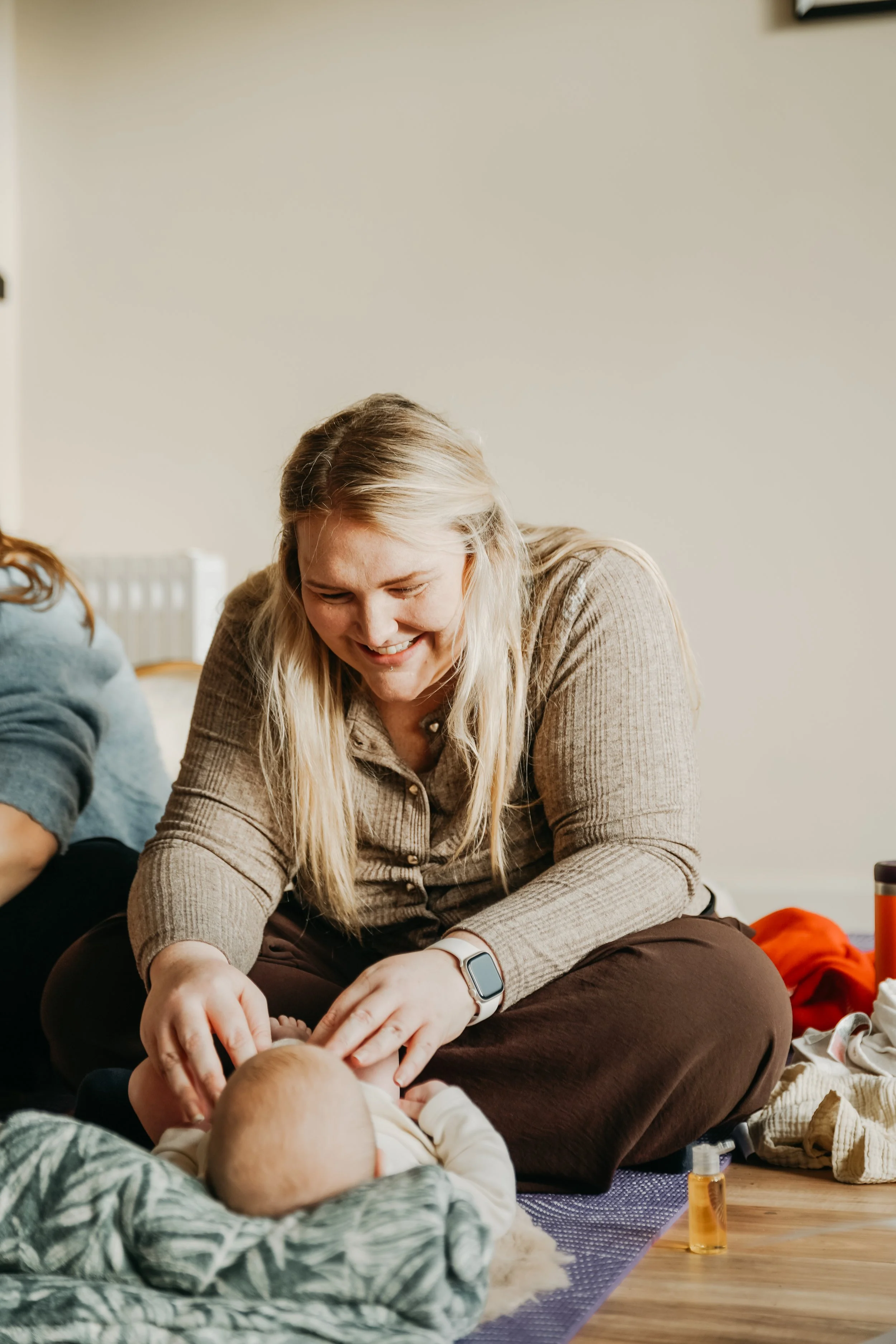 A woman with long blonde hair smiling while sitting on the floor, tending to a baby, with another person partially visible on her left, in a cozy indoor setting.