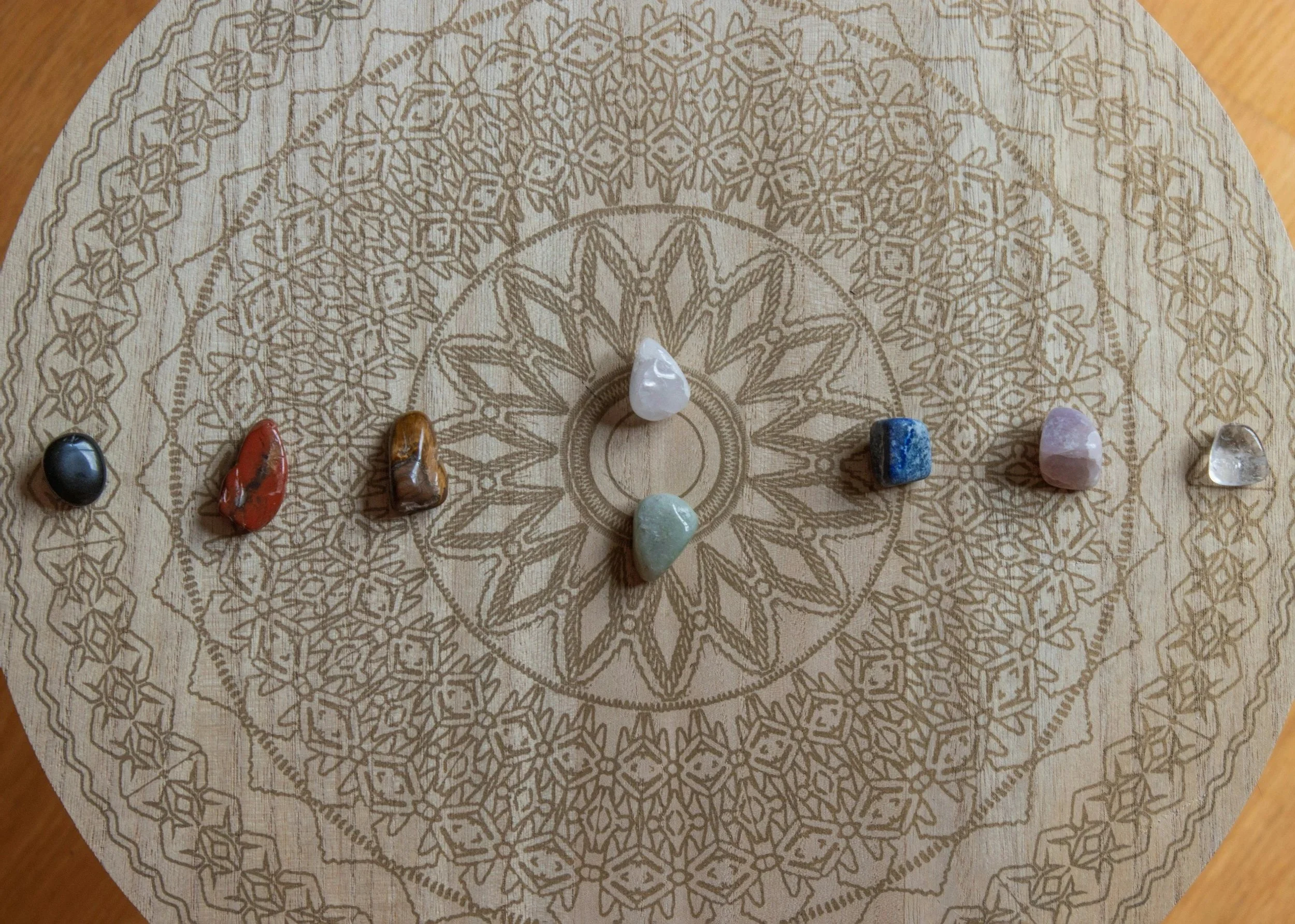 A circular wooden board with intricate mandala-inspired geometric patterns engraved on it. Several small polished gemstones in various colors are arranged at the lower part of the board.
